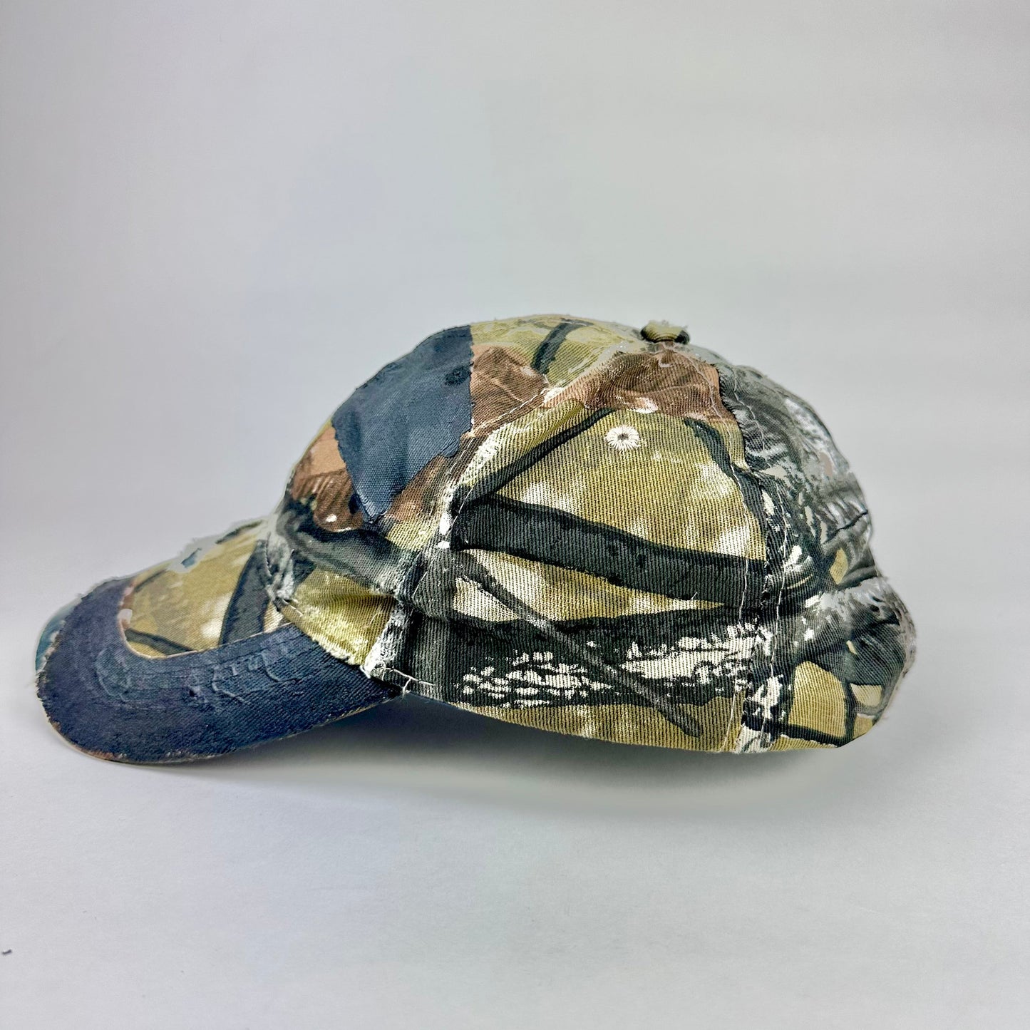 Still life left side of ASTROBOI* x Lowe’s MVP brown RealTree camouflage hat with resin-sealed rhinestone logo, black waxed tattoo print, adjustable strap, custom NYC piece