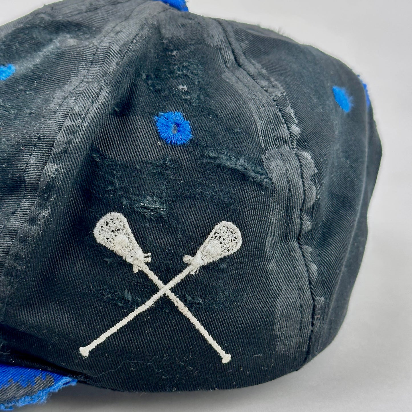 Black cap with blue accents and white lacrosse stick design on a light gray background