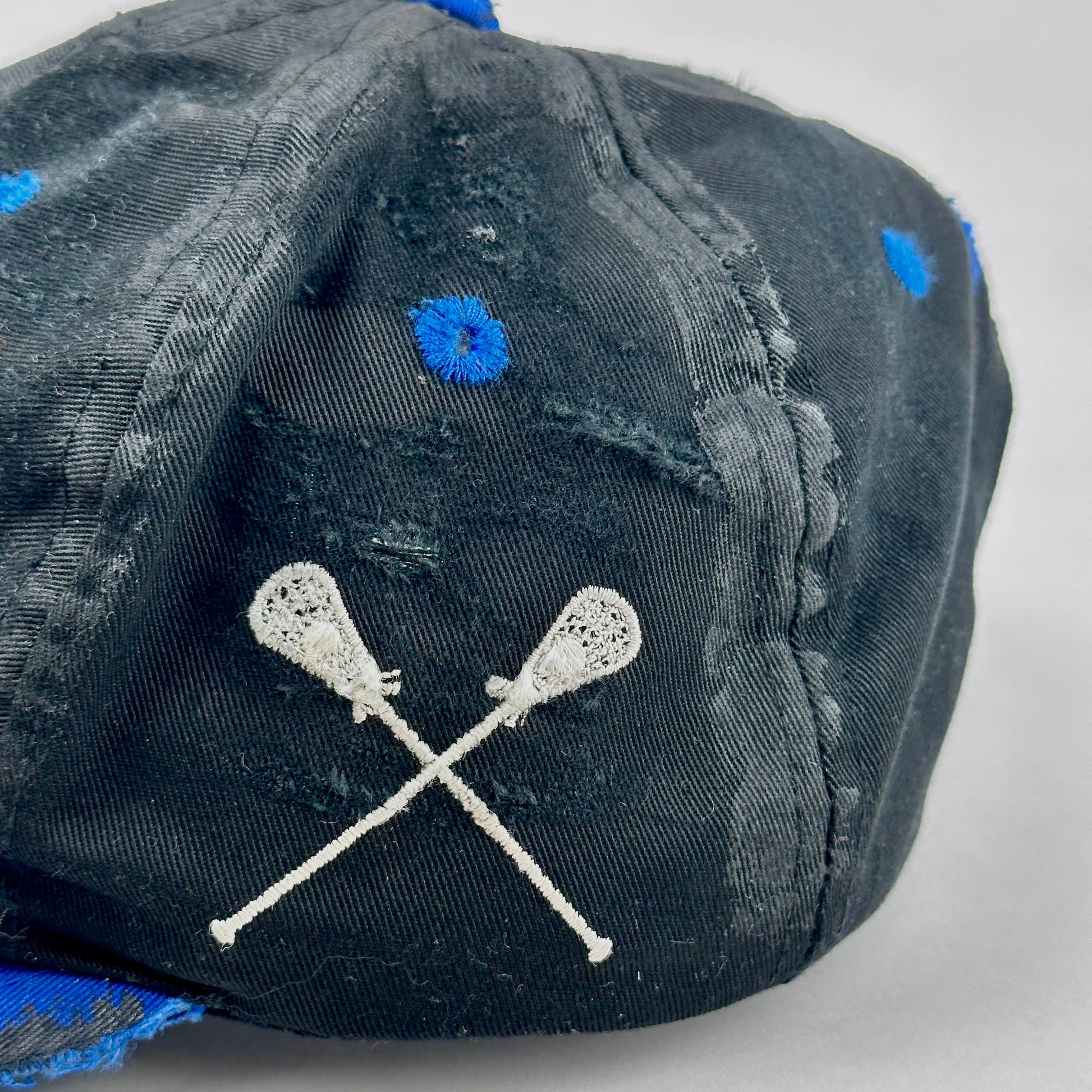Black cap with blue accents and white lacrosse stick design on a light gray background