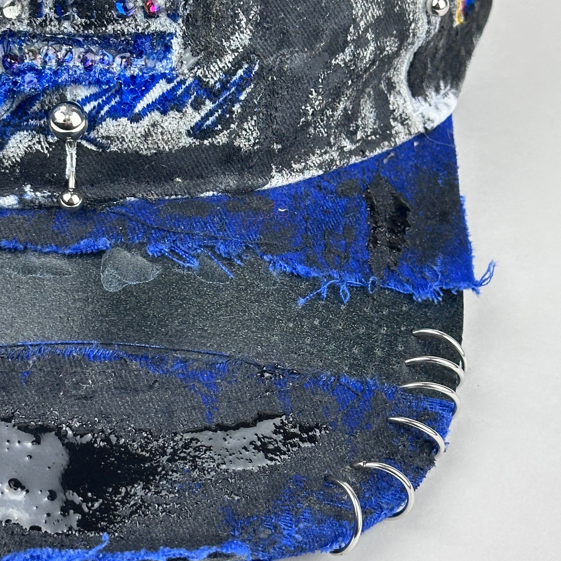 Right side of visor piercing detail on Blue and white ASTROBOI* NAPA hat with rhinestone-covered logo sealed in clear resin, stainless steel piercings, and a deconstructed visor, handcrafted in NYC.