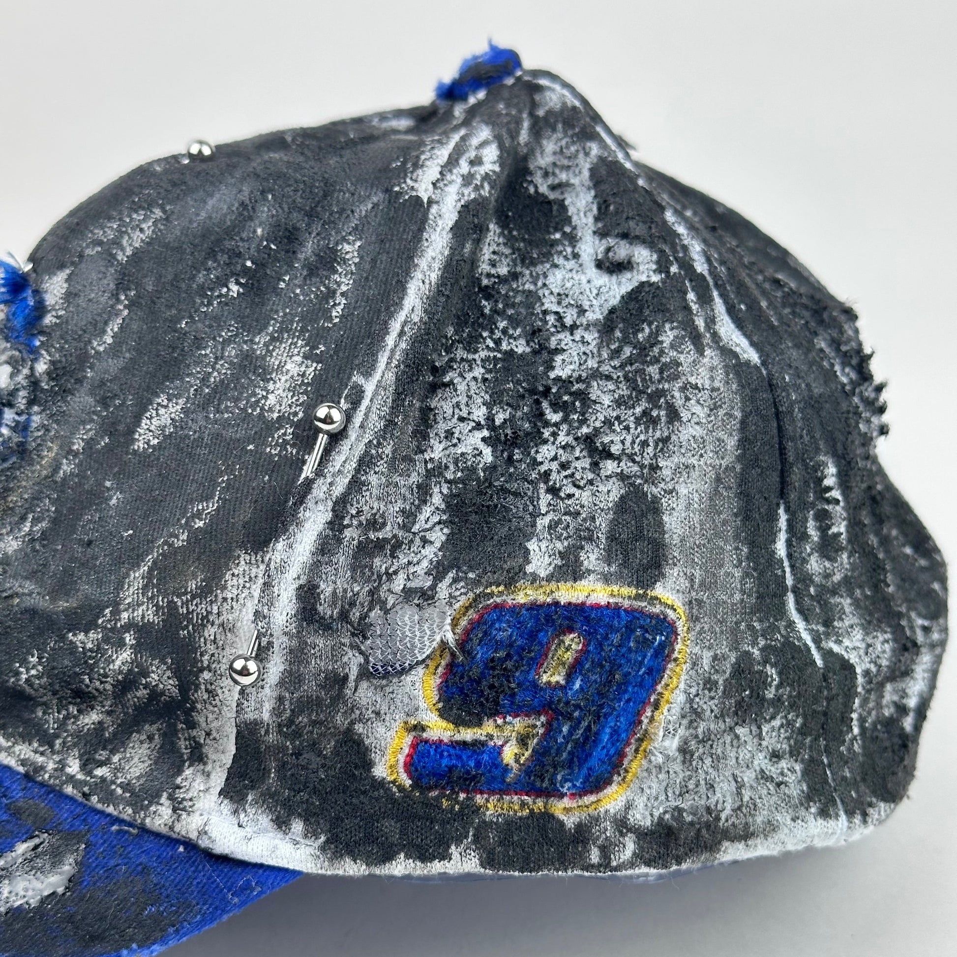Left side detail of embroidered 9 on Blue and white ASTROBOI* NAPA hat with rhinestone-covered logo sealed in clear resin, stainless steel piercings, and a deconstructed visor, handcrafted in NYC.