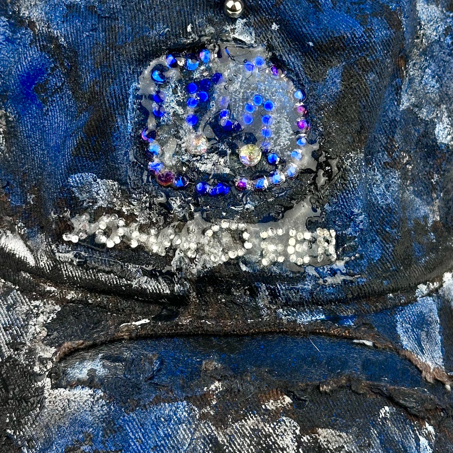 Close up od logo on ASTROBOI* x John Deere black and blue distressed trucker hat with rhinestone resin logo and stainless steel piercings.
