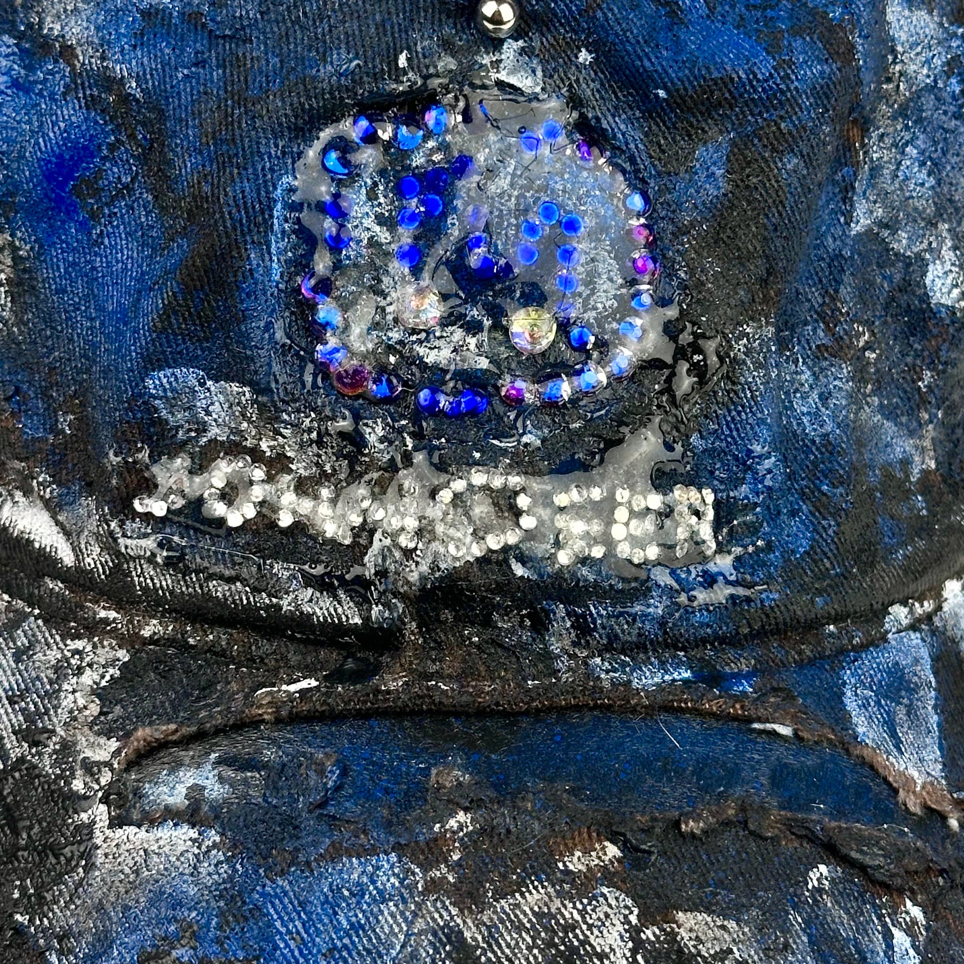 Close up od logo on ASTROBOI* x John Deere black and blue distressed trucker hat with rhinestone resin logo and stainless steel piercings.