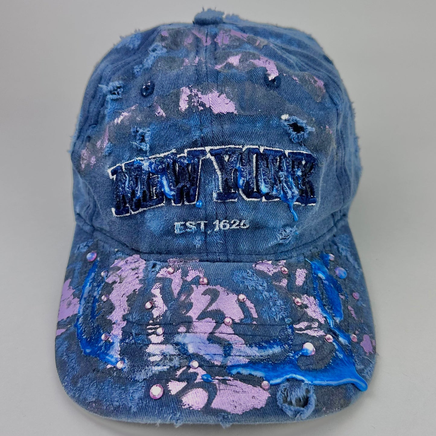 Front image of ASTROBOI* 'Twilight Haze' distressed blue resin hat with New York logo, lavender metallic paint treatment, oozing resin effect, and matching lavender rhinestone embellishments, handmade in NYC.