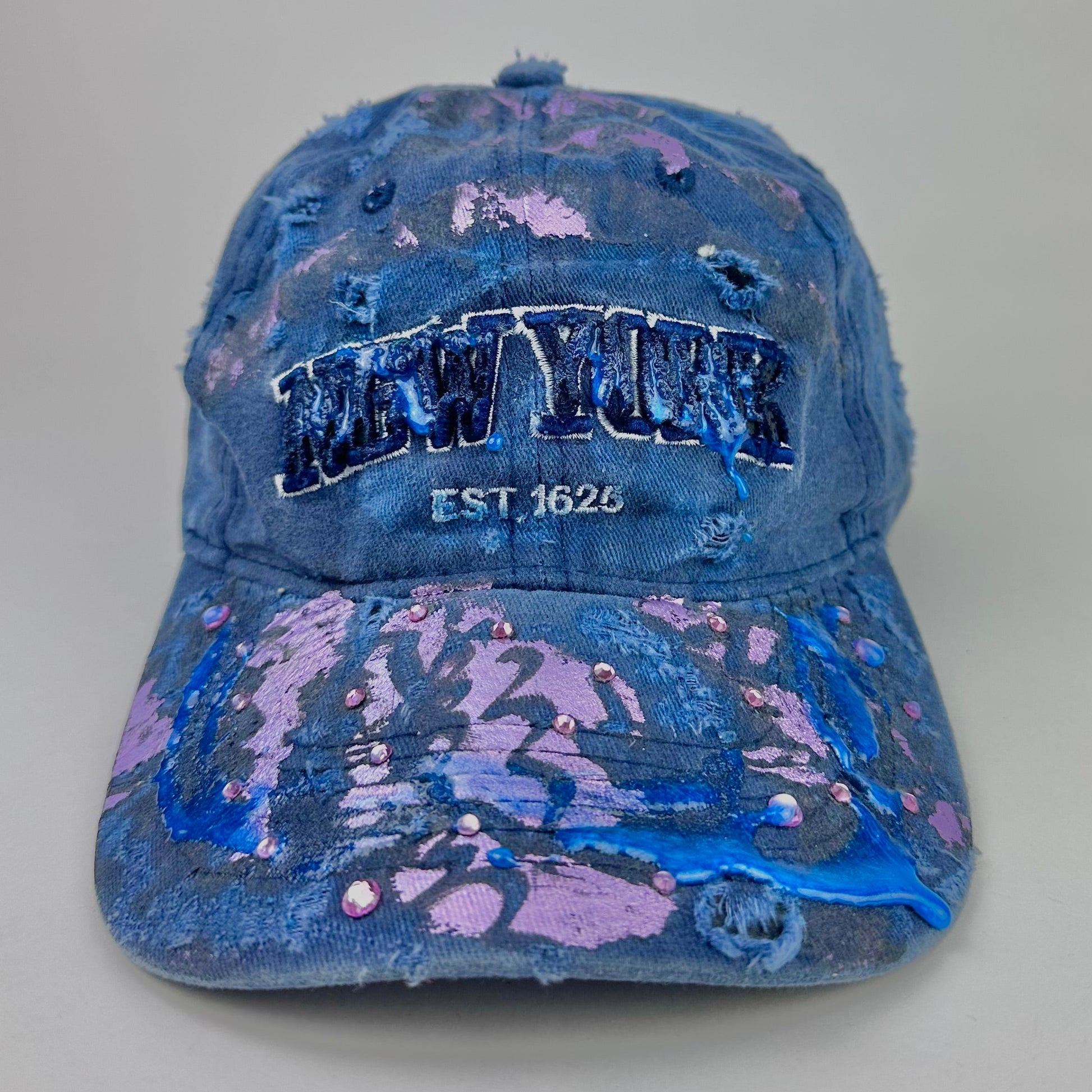 Visor detail on ASTROBOI* 'Twilight Haze' distressed blue resin hat with New York logo, lavender metallic paint treatment, oozing resin effect, and matching lavender rhinestone embellishments, handmade in NYC.