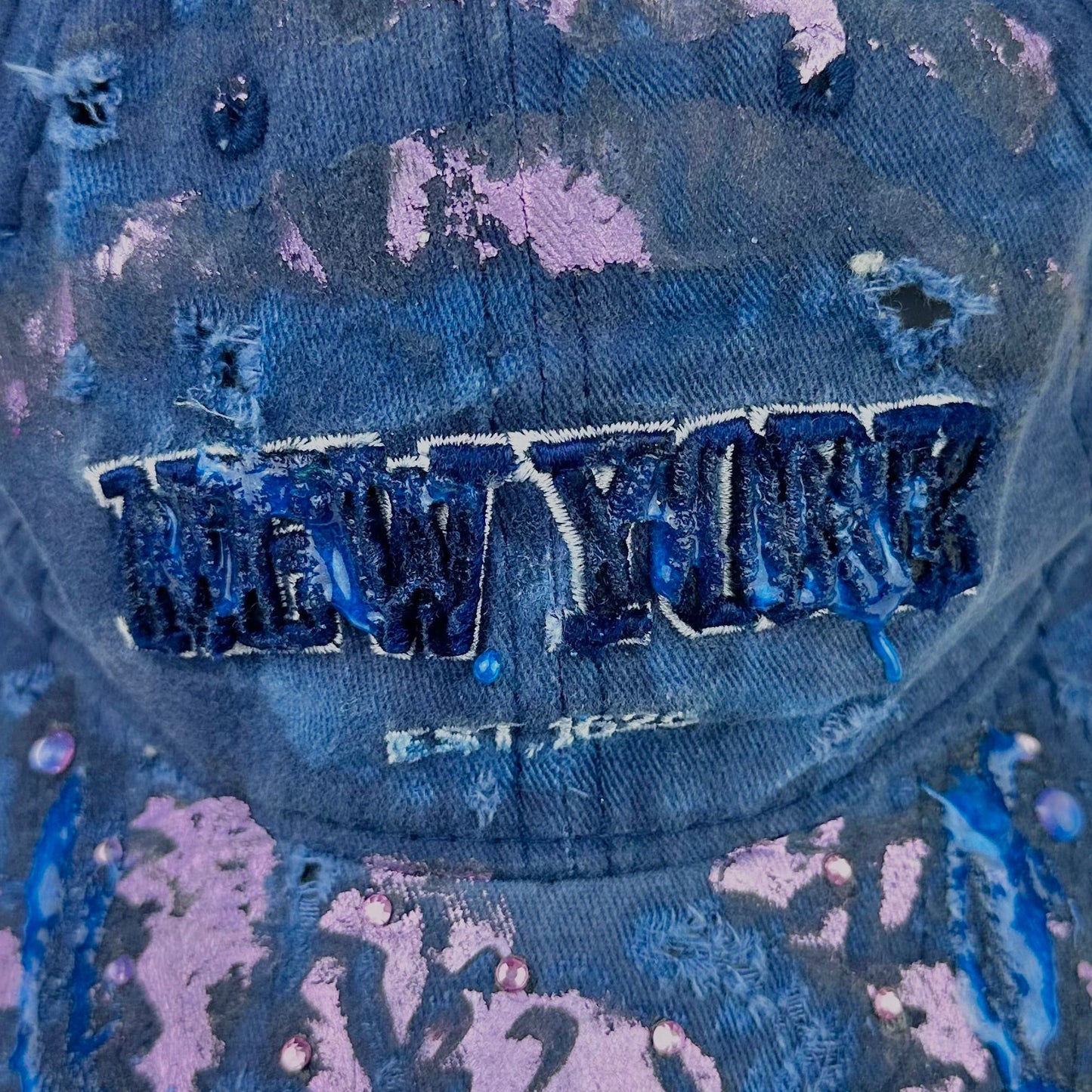 Logo detail on ASTROBOI* 'Twilight Haze' distressed blue resin hat with New York logo, lavender metallic paint treatment, oozing resin effect, and matching lavender rhinestone embellishments, handmade in NYC.