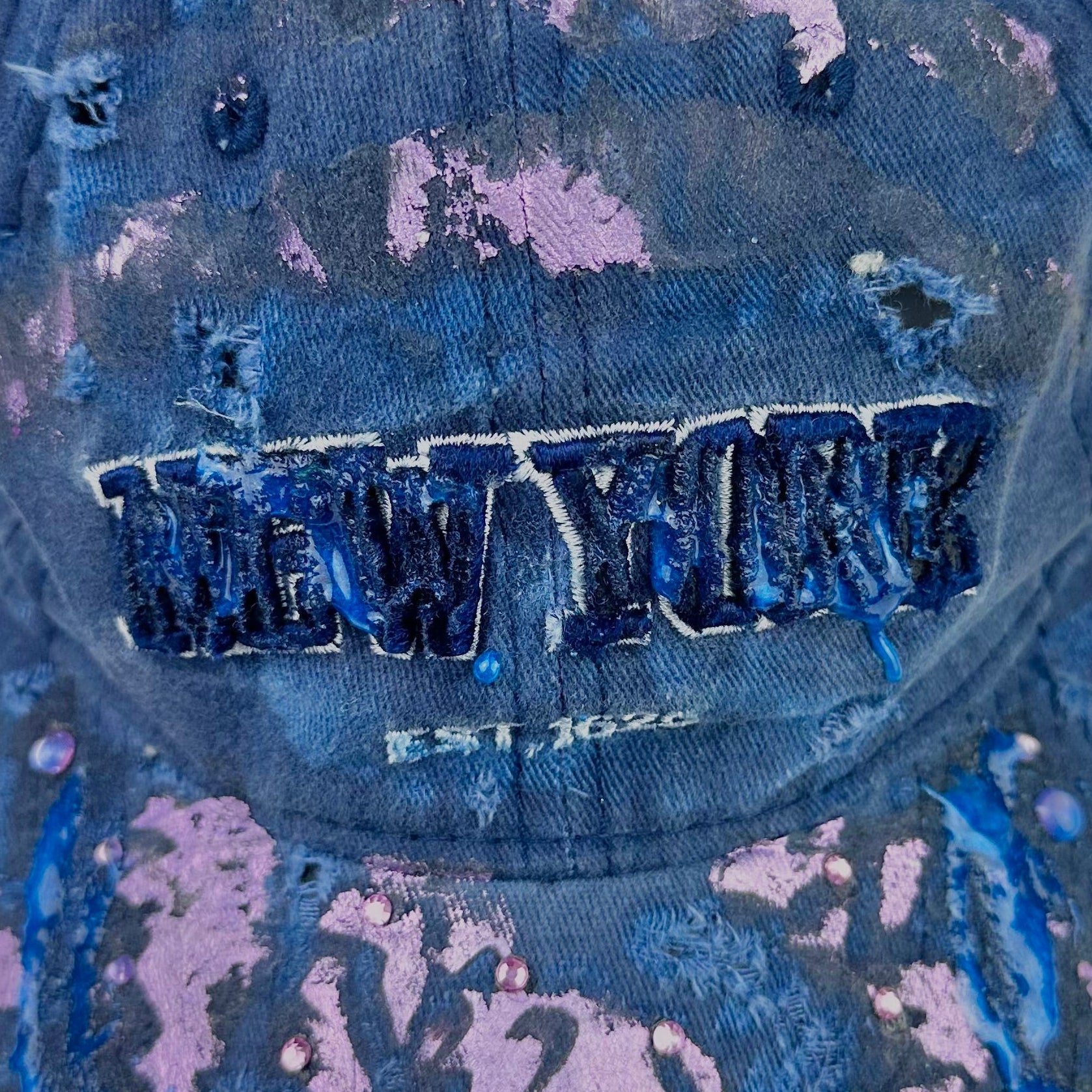 Logo detail on ASTROBOI* 'Twilight Haze' distressed blue resin hat with New York logo, lavender metallic paint treatment, oozing resin effect, and matching lavender rhinestone embellishments, handmade in NYC.