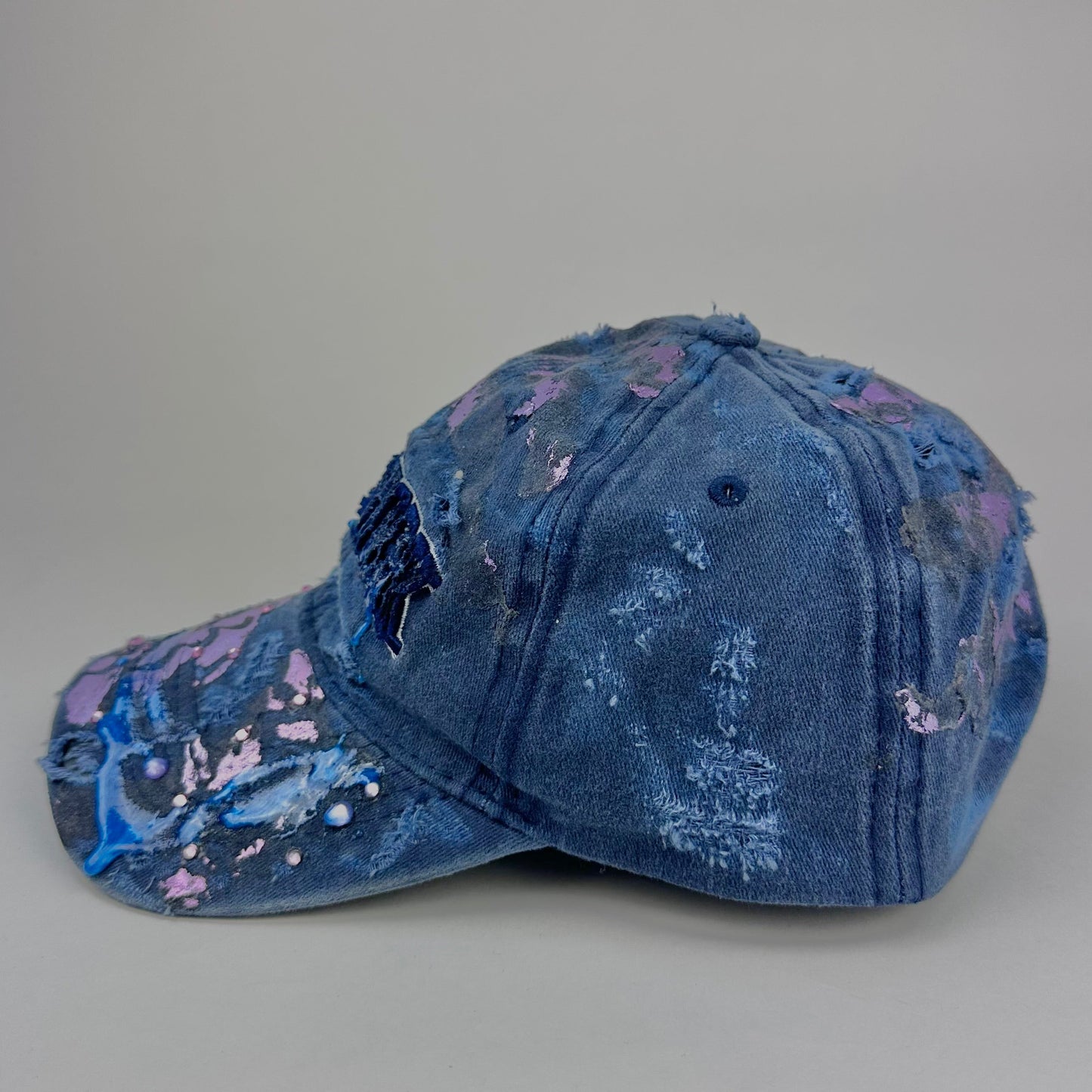 Left side of ASTROBOI* 'Twilight Haze' distressed blue resin hat with New York logo, lavender metallic paint treatment, oozing resin effect, and matching lavender rhinestone embellishments, handmade in NYC.