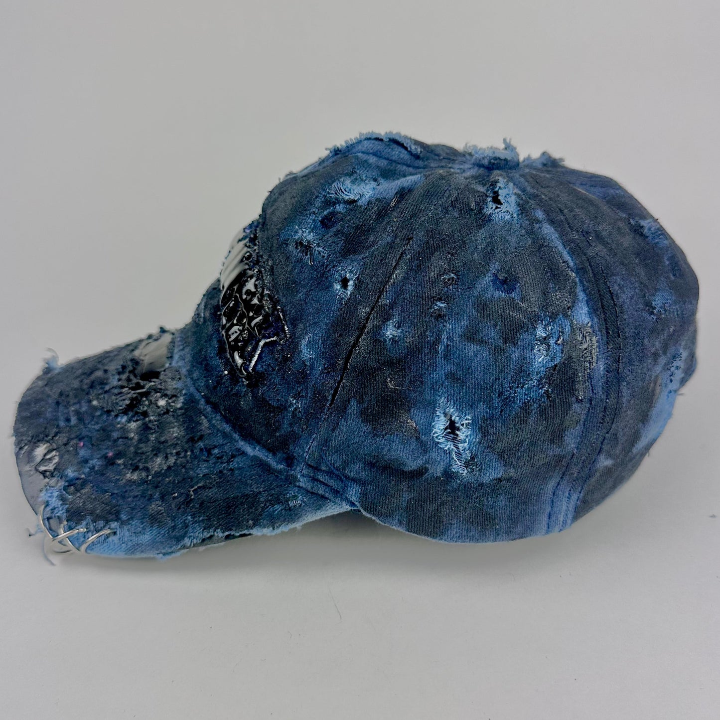 Left side of ASTROBOI* 'Feeling Blue' distressed blue resin-coated hat with hand-painted black shading, metal piercings on the right front, and New York embroidery, handmade in NYC.