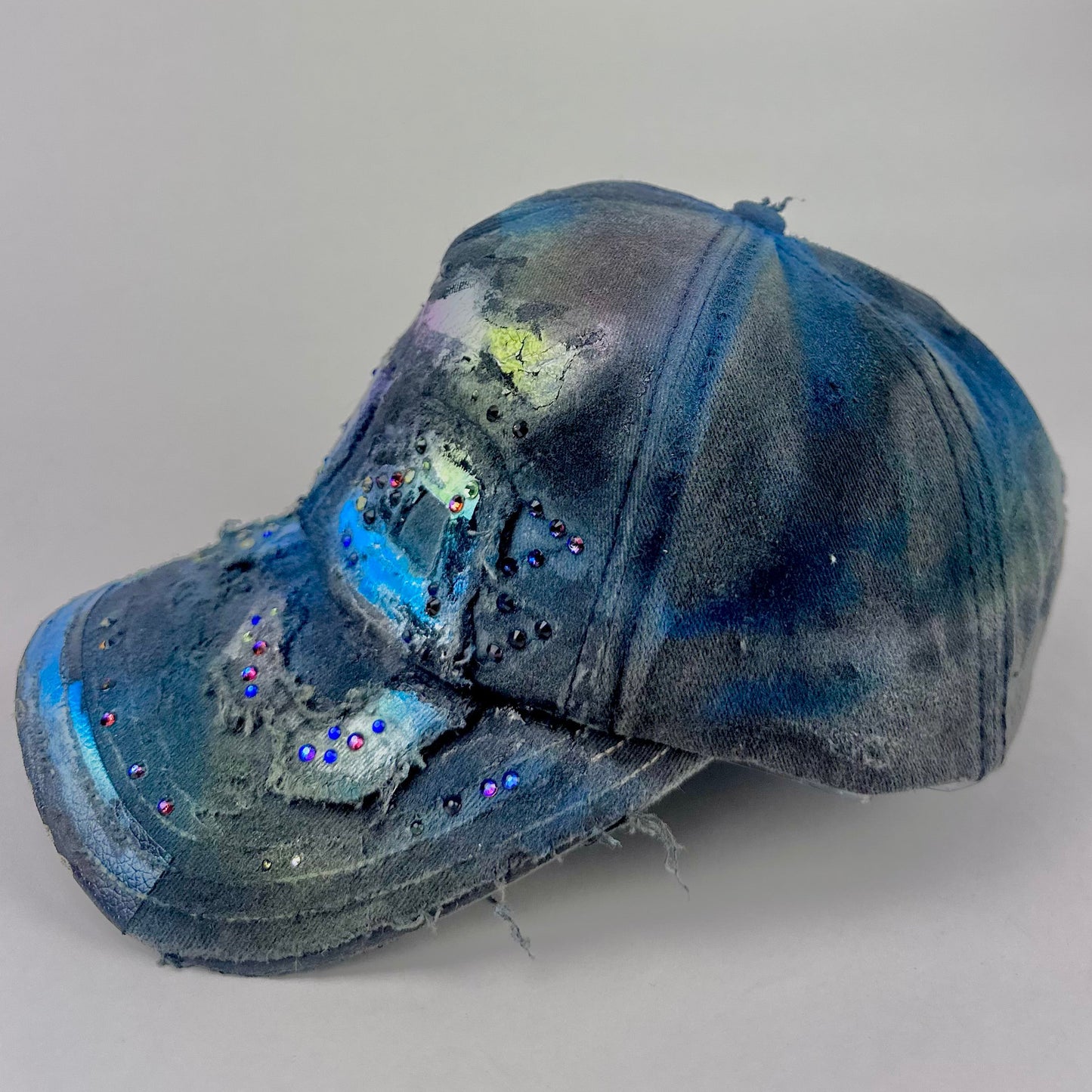 Left side of ASTROBOI* 'Tropical 79' upcycled Y2K distressed hat featuring rainbow paint design, multicolored rhinestone embellishments, and hand-painted black shading for contrast, on a neutral background.