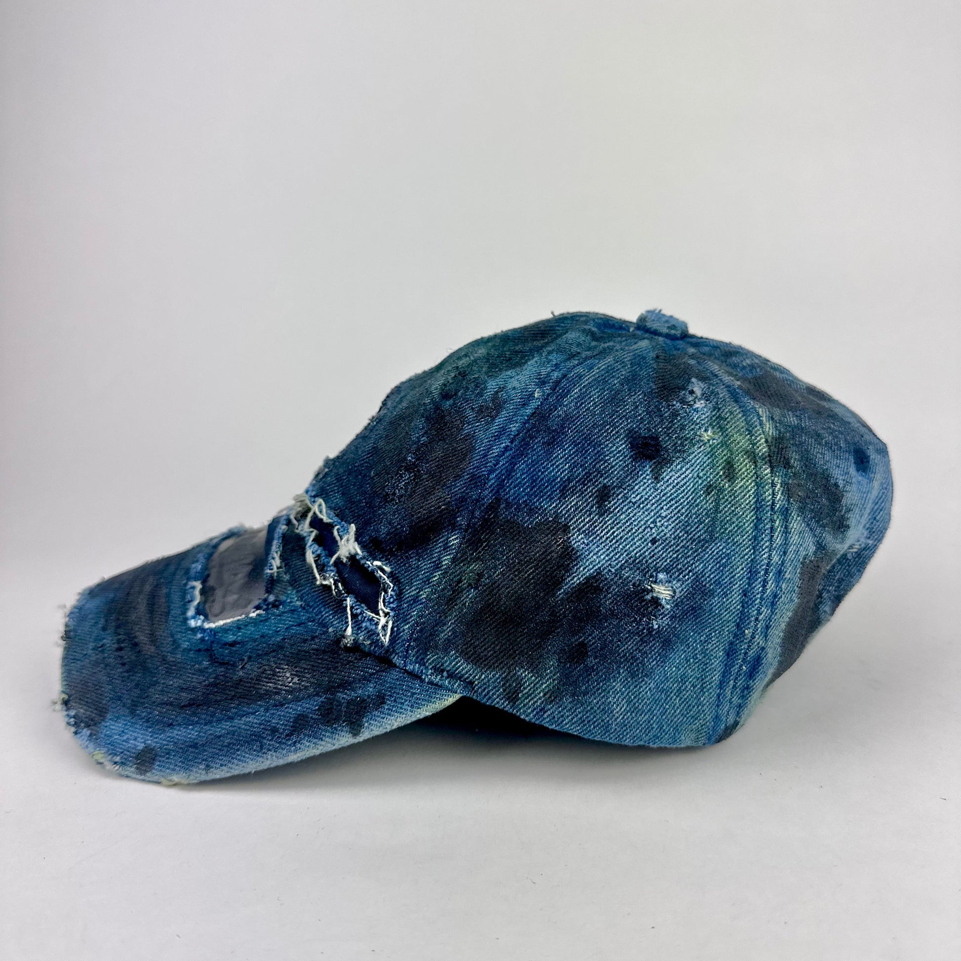 Left side of ASTROBOI* 'C' deconstructed black and blue faded denim hat with hand-painted blue-green shading, raw crown cut-outs, and embossed C logo at center front, customized in NYC.