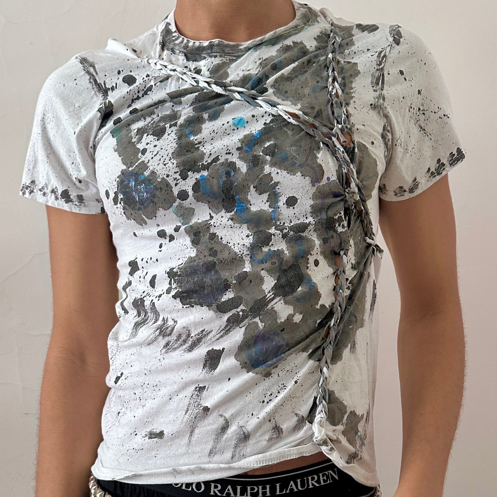 Close-up of ASTROBOI* x Gildan 'Hidden Messages' showing multicolor blue scribble paint, white watercolor ink splatter layering, and front braid detail on white cotton fabric.