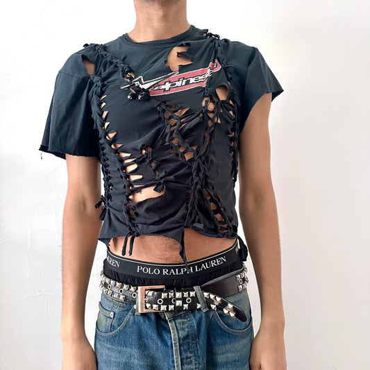 ASTROBOI* x Alpinestars black braided cut-out t-shirt with hand-braided detailing, ripped construction, asymmetric sleeves, uneven hem, and Alpinestars logo at front neck, front view.