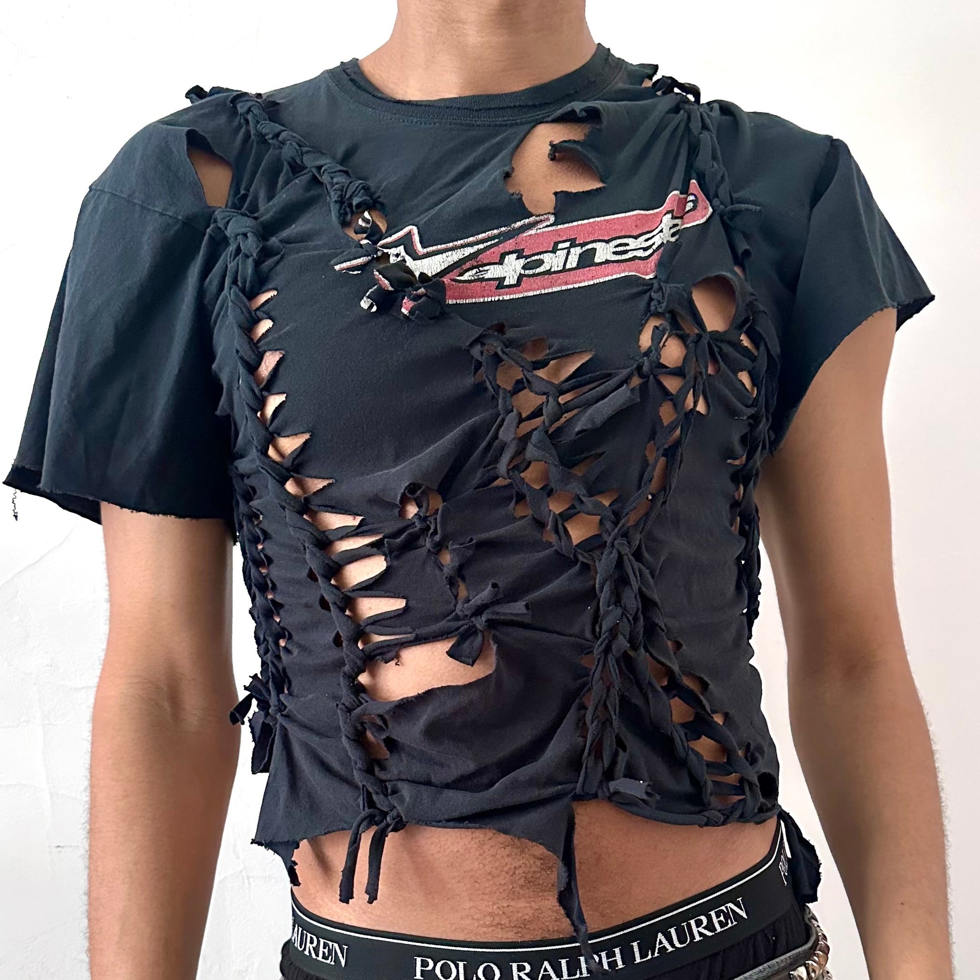 Close-up of ASTROBOI* x Alpinestars tee showing hand-braided construction, ripped detailing, and Alpinestars logo on black fabric.