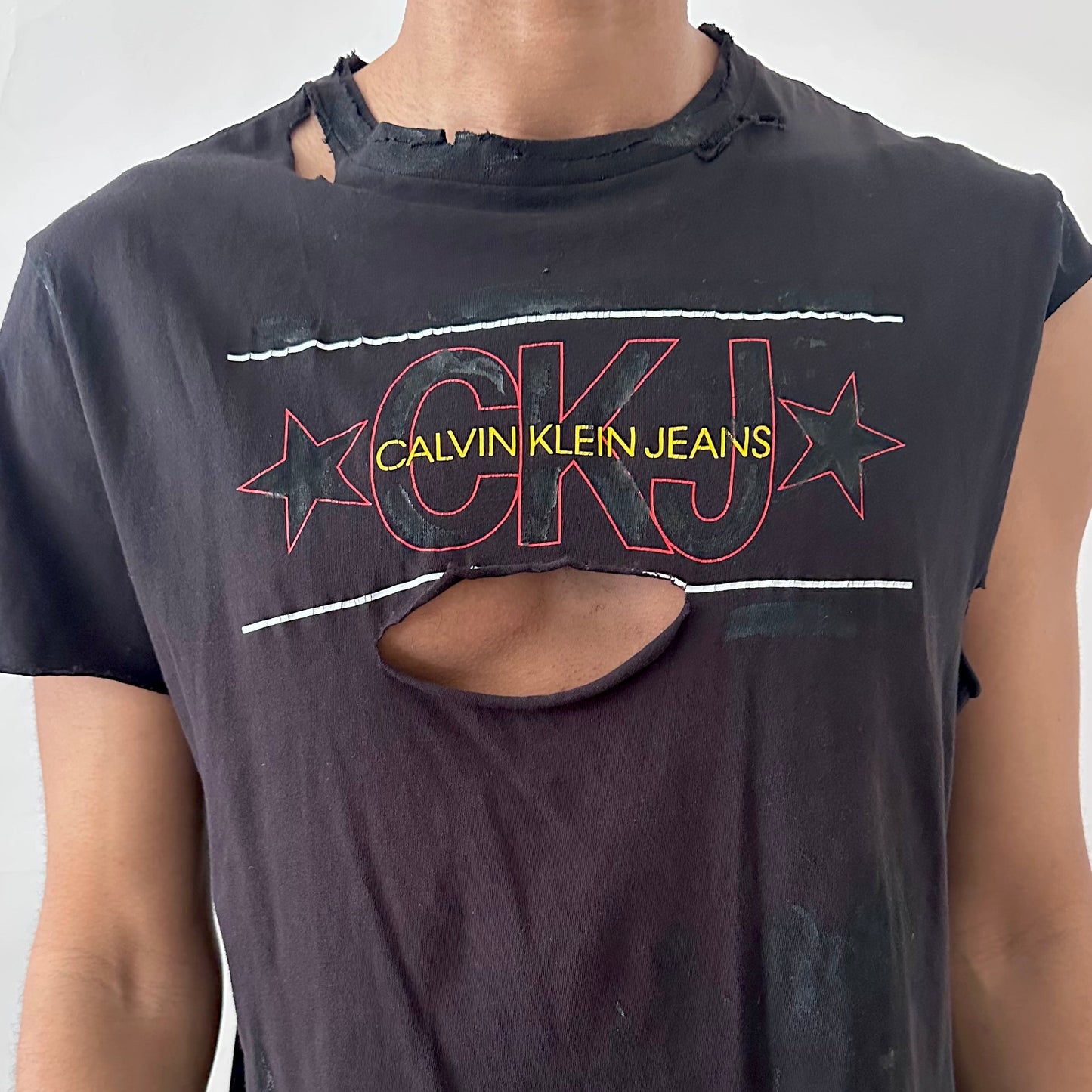 Close-up of ASTROBOI* x CK Jeans asymmetric logo tee showing CK Jeans chest logo and deconstructed asymmetric sleeve detail on black fabric.
