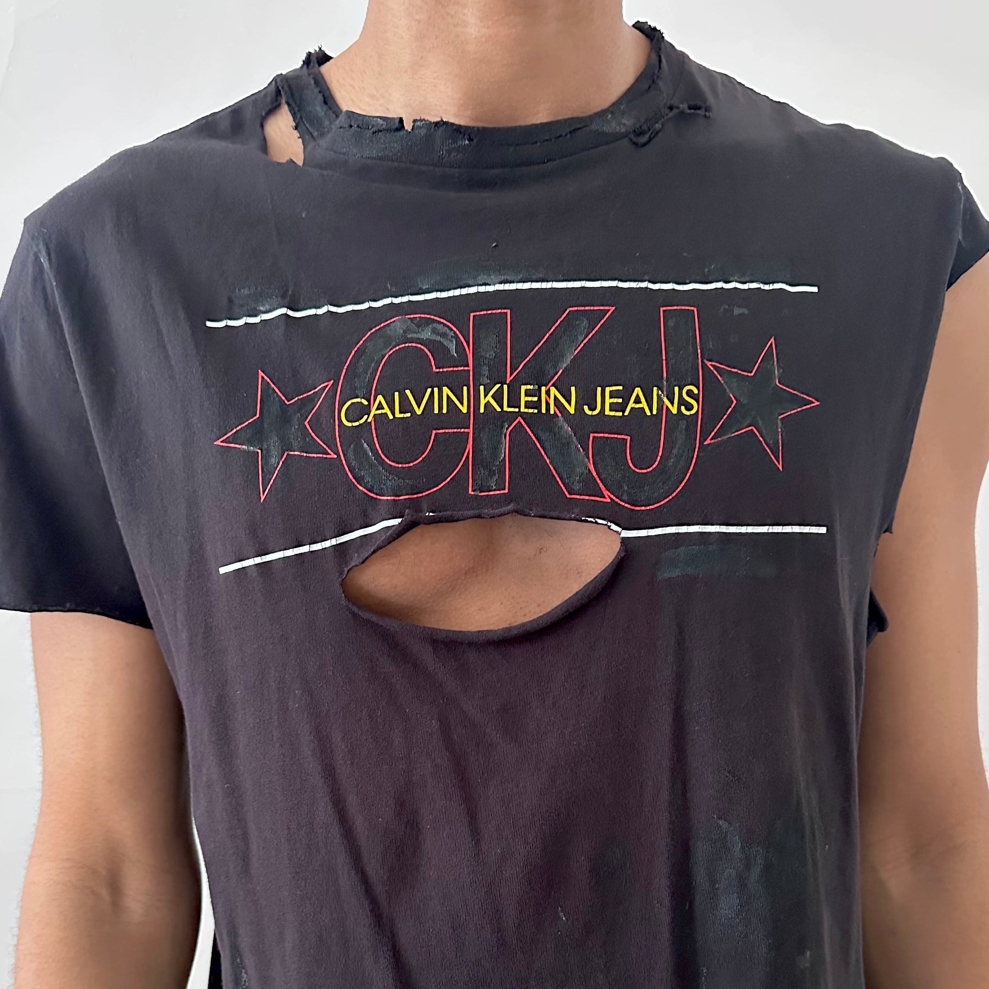 Close-up of ASTROBOI* x CK Jeans asymmetric logo tee showing CK Jeans chest logo and deconstructed asymmetric sleeve detail on black fabric.