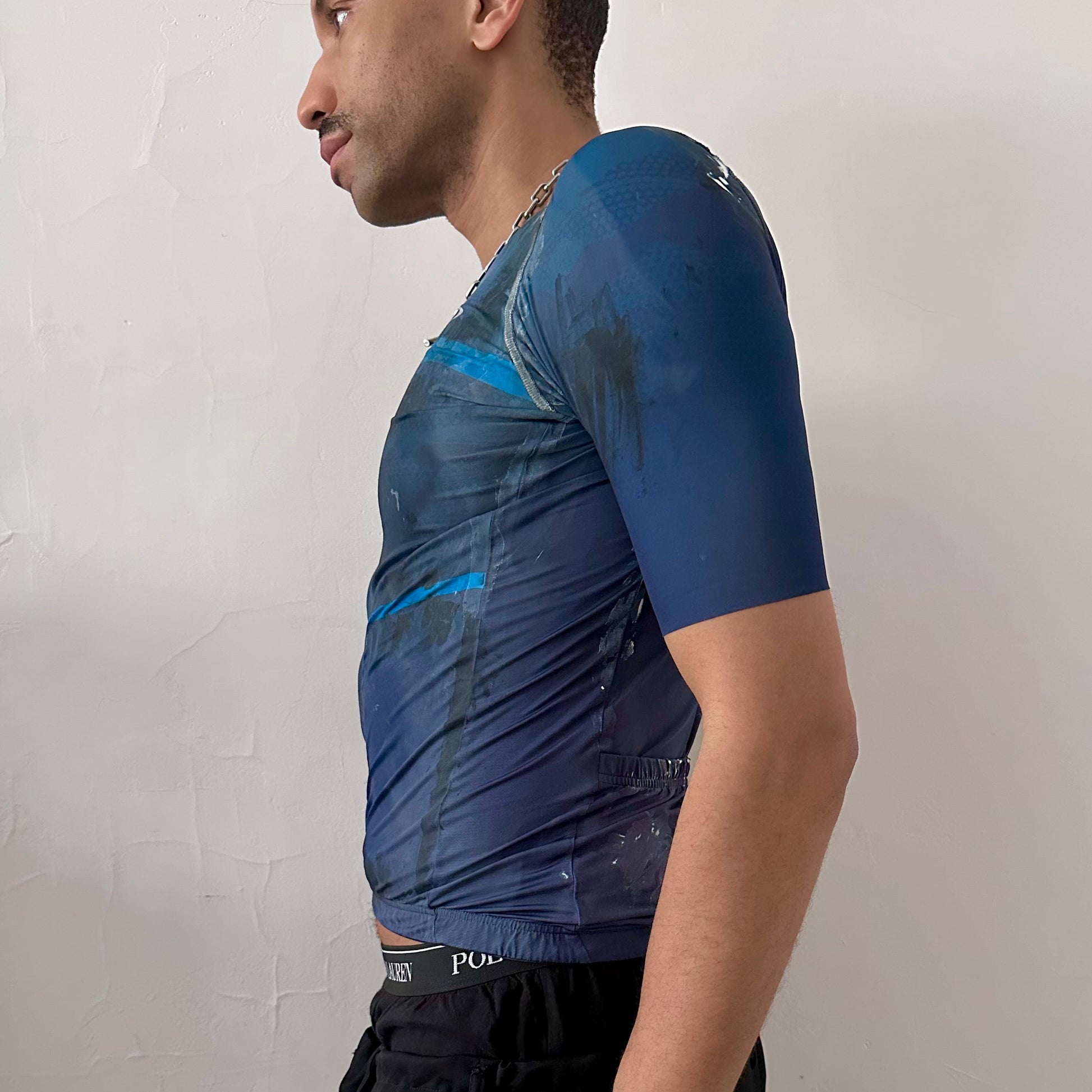 Left side detail of ASTROBOI* x Oakley cycling jersey showing hand-applied painterly pattern and zipper closure on blue and black fabric.