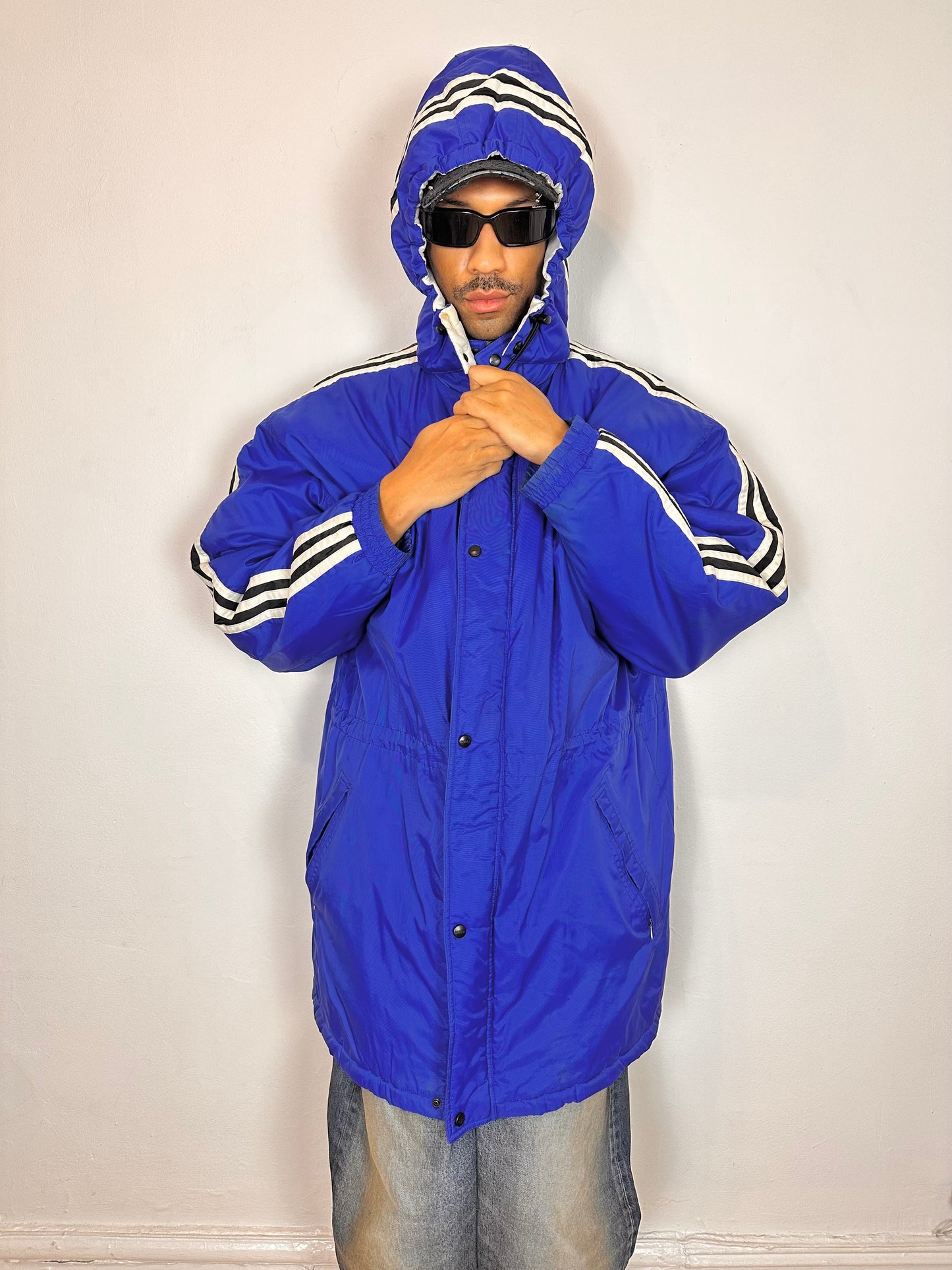 Front styled view of Vintage Adidas blue oversized winter puffer coat with detachable hood, white stripes, and long insulated silhouette, size XXL.