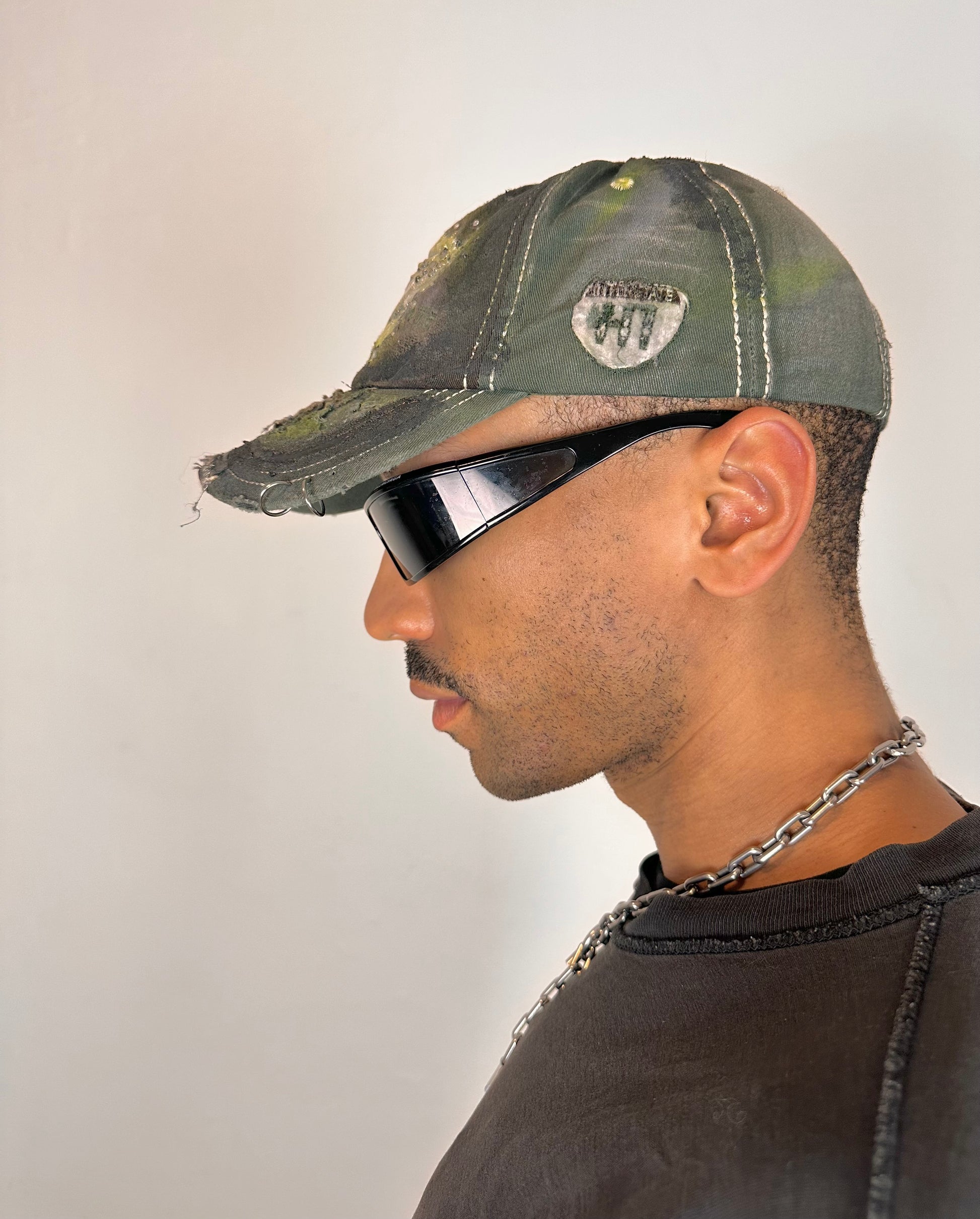 Left side of “Green and brown distressed ASTROBOI* Hawaii hat with rhinestones, metallic paint, and sterling-steel piercings, customized in NYC.”
