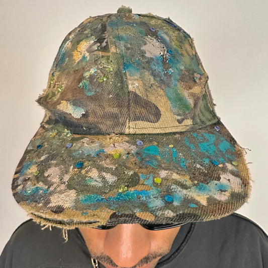 Front of ASTROBOI* distressed brown camouflage cap with metallic silver and blue paint and tonal rhinestone accents, adjustable velcro back.