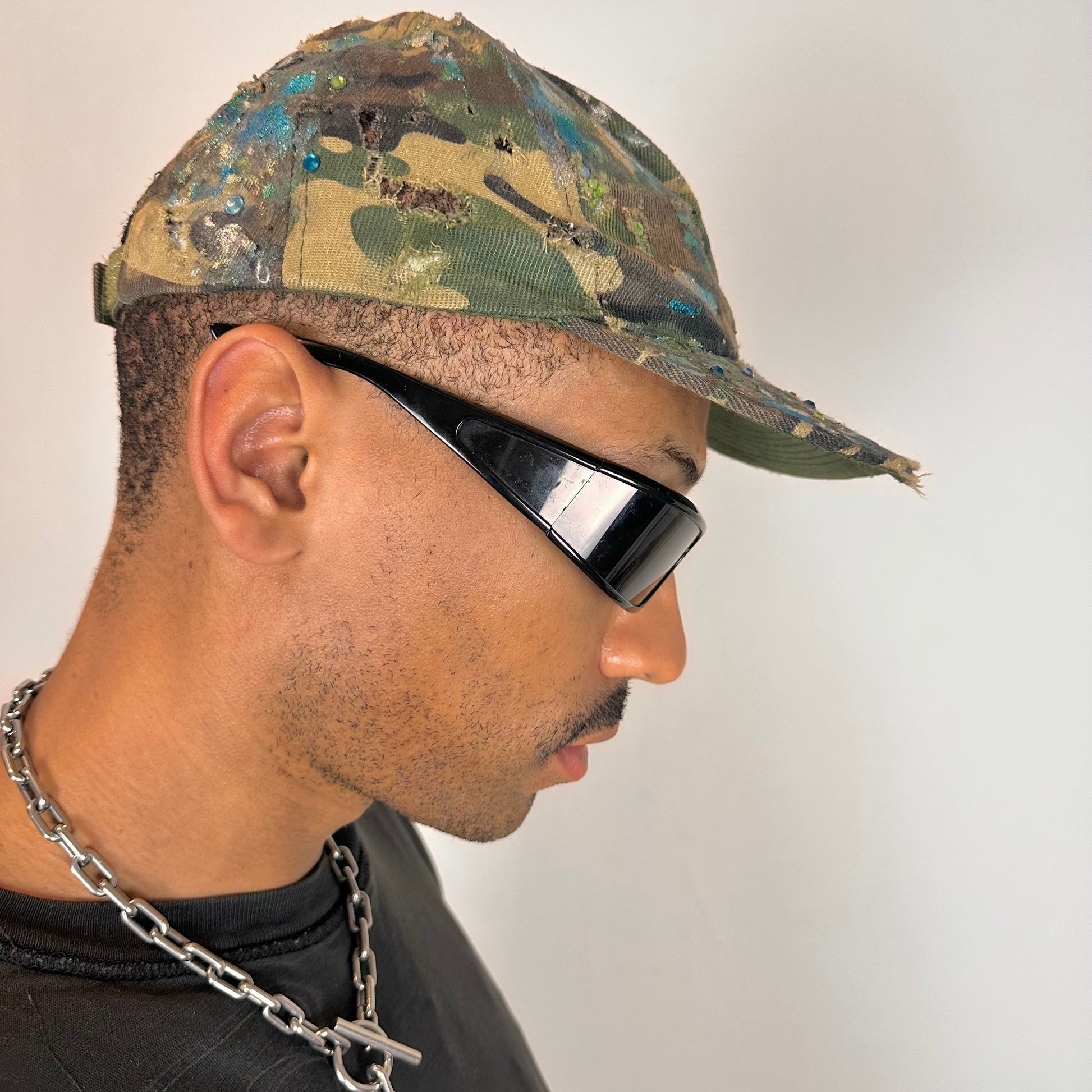 Right side of ASTROBOI* distressed brown camouflage cap with metallic silver and blue paint and tonal rhinestone accents, adjustable velcro back.