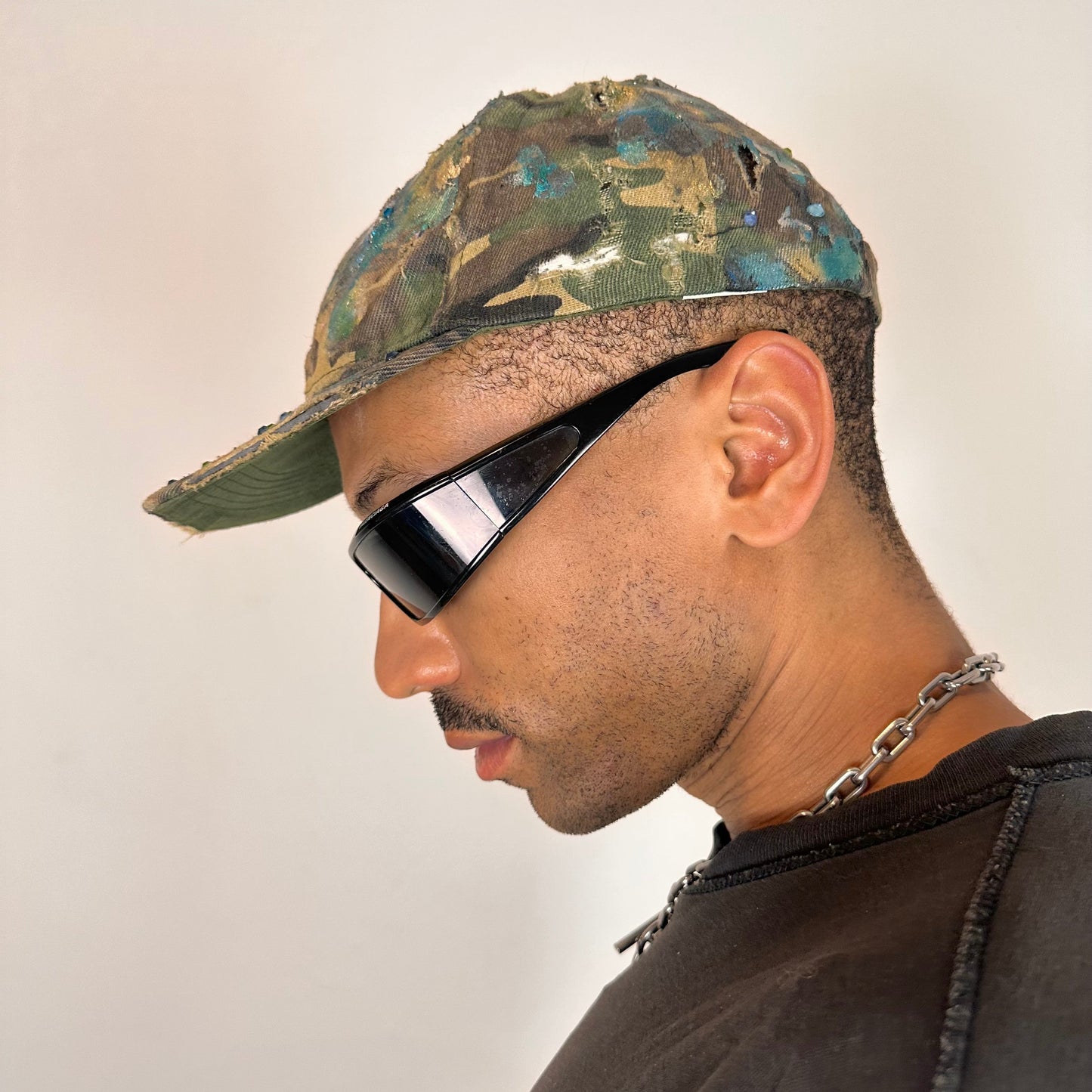 Left side of ASTROBOI* distressed brown camouflage cap with metallic silver and blue paint and tonal rhinestone accents, adjustable velcro back.