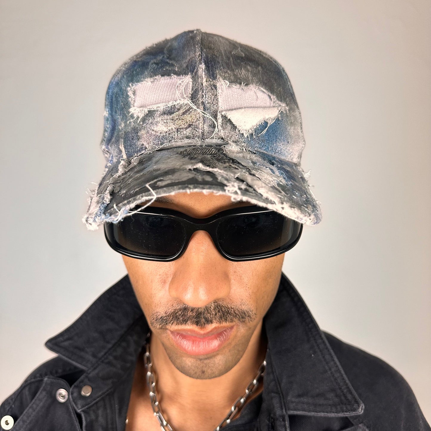 Overview of ASTROBOI* Margiela blue deconstructed hat with distressed tears, airbrushed paint, denim scraps, silver staples and resin-coated visor.