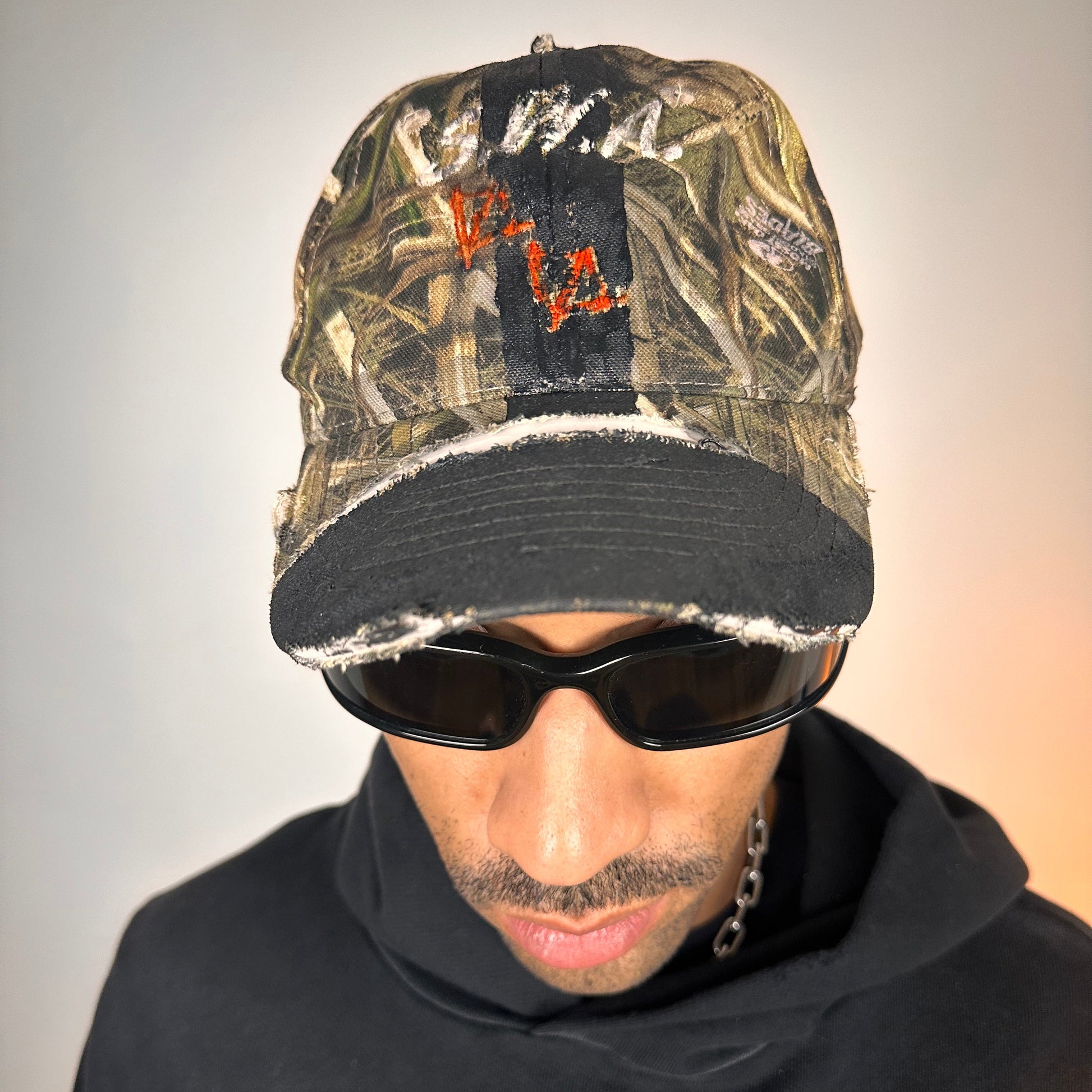 Front of ASTROBOI* x Mossy Oak brown RealTree camouflage tattoo hat with SWA embroidery, black painted distressing, youth size, custom NYC piece