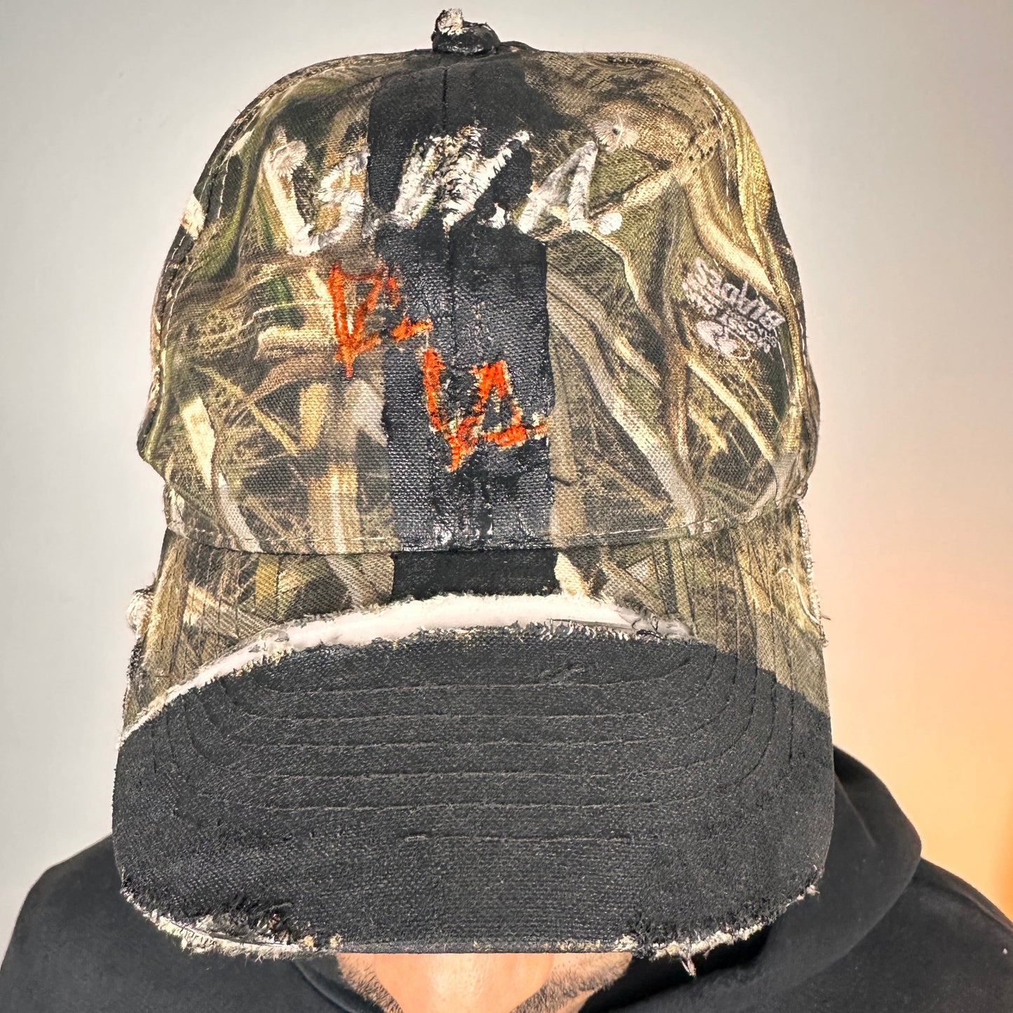 Close up of ASTROBOI* x Mossy Oak brown RealTree camouflage tattoo hat with SWA embroidery, black painted distressing, youth size, custom NYC piece