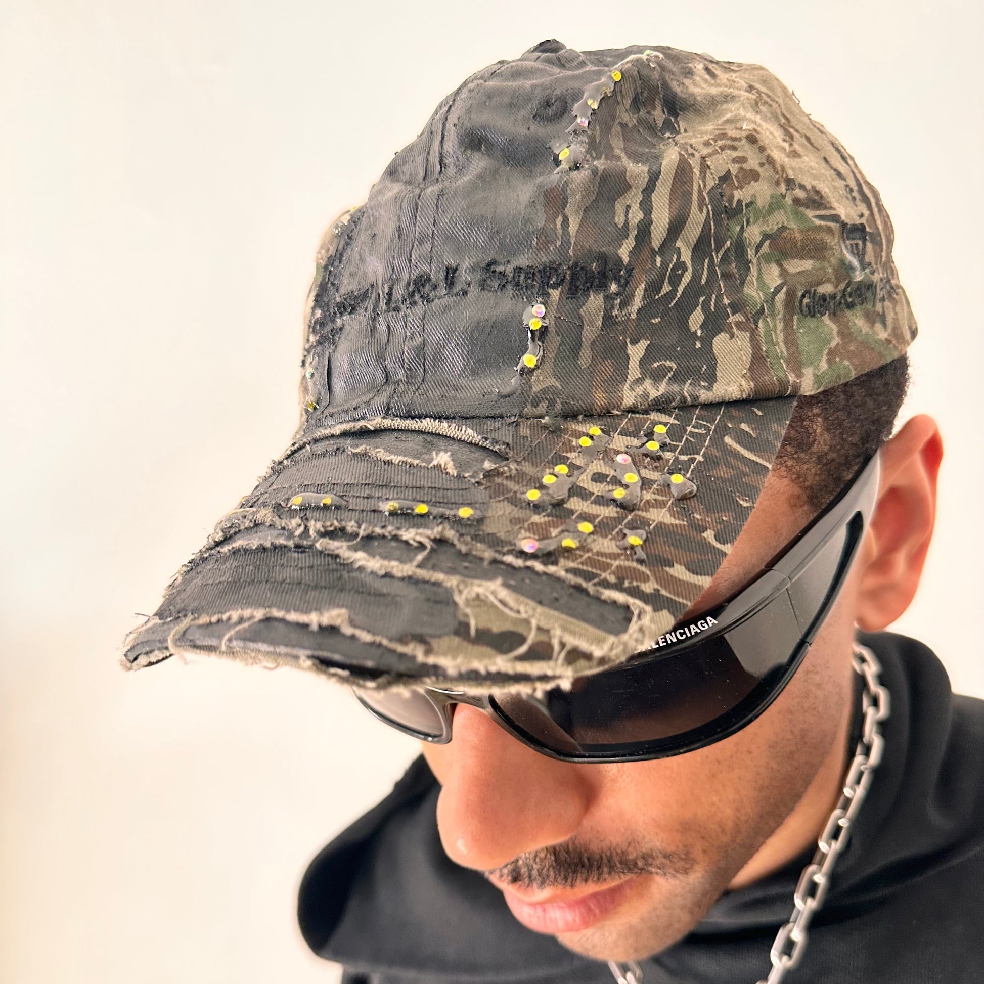 Front left side of ASTROBOI* L&L Supply green RealTree camo utility hat with purple and green rhinestones sealed in resin and black tattoo print detailing