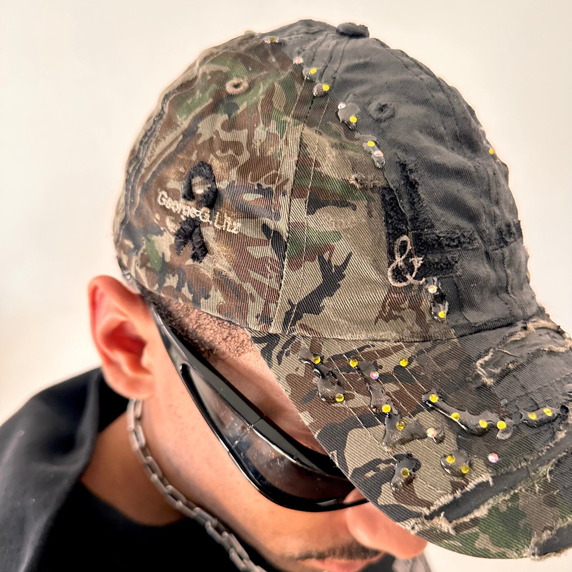 Front right detail on ASTROBOI* L&L Supply green RealTree camo utility hat with purple and green rhinestones sealed in resin and black tattoo print detailing
