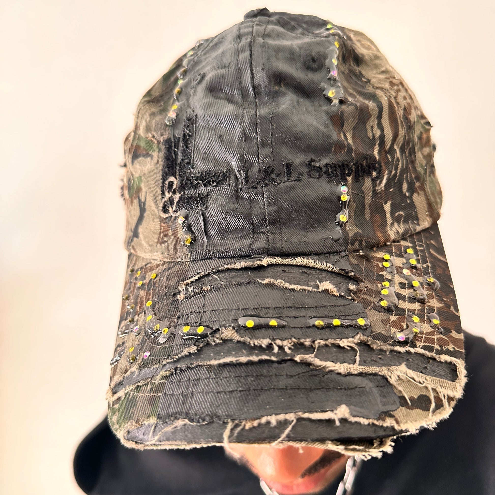 Cover shot of ASTROBOI* L&L Supply green RealTree camo utility hat with purple and green rhinestones sealed in resin and black tattoo print detailing