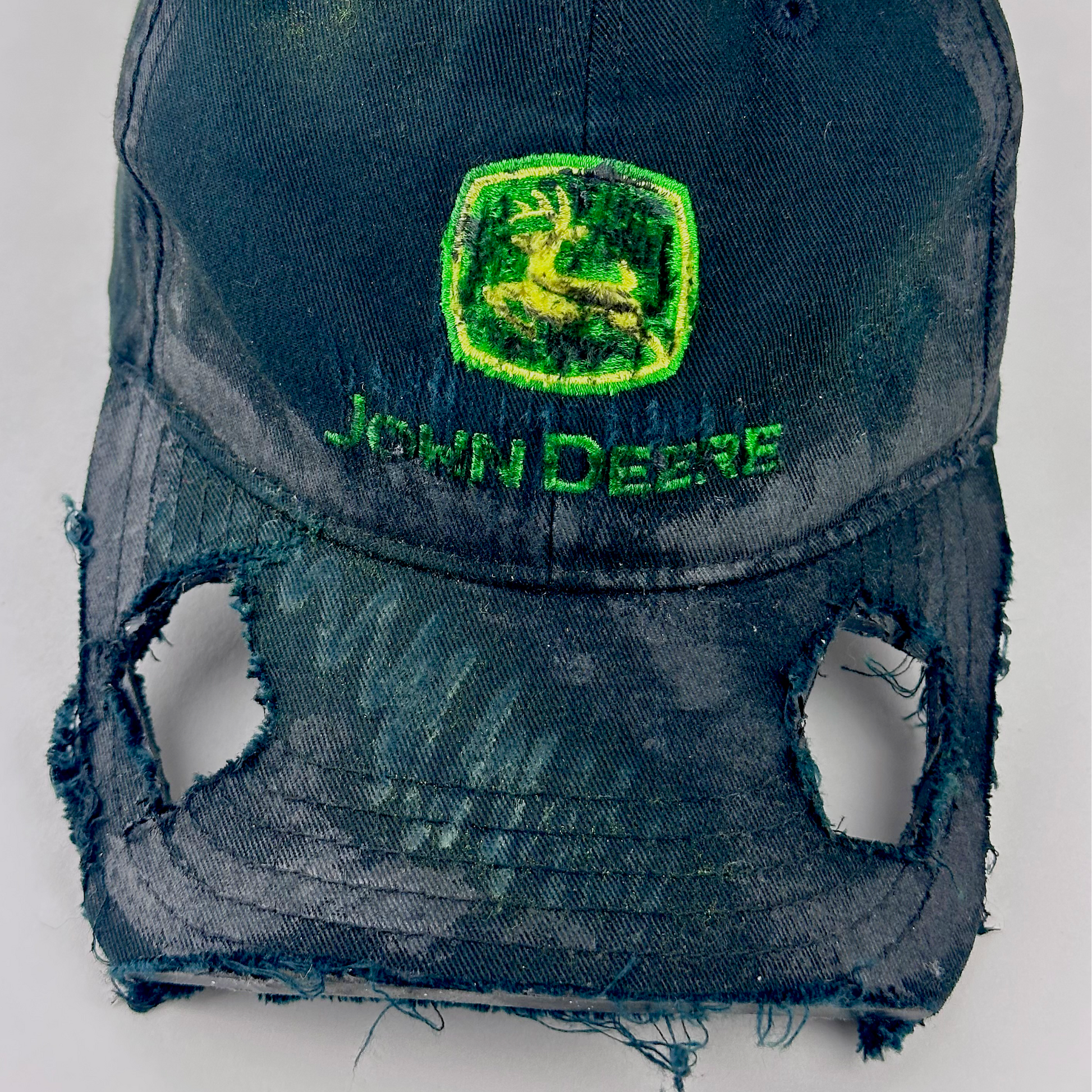 Close-up of ASTROBOI* x John Deere 'Hyland' showing John Deere green and yellow logo patch and cut-out visor detail with custom paint treatment on black distressed fabric.