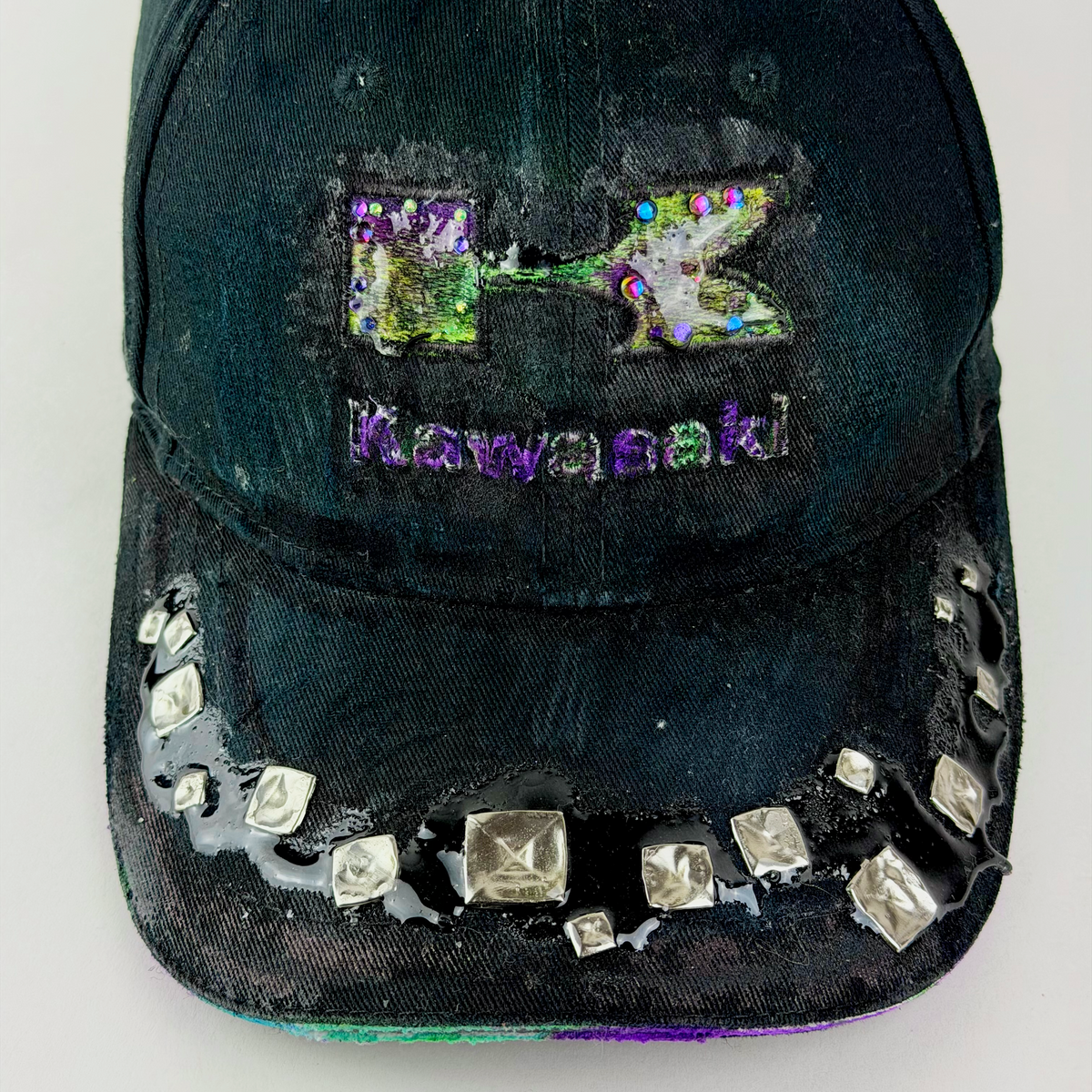A distressed black baseball cap featuring a holographic 'K' logo and 'Kawasaki' text on the front, embellished with silver studs along the brim, set against a plain light background.