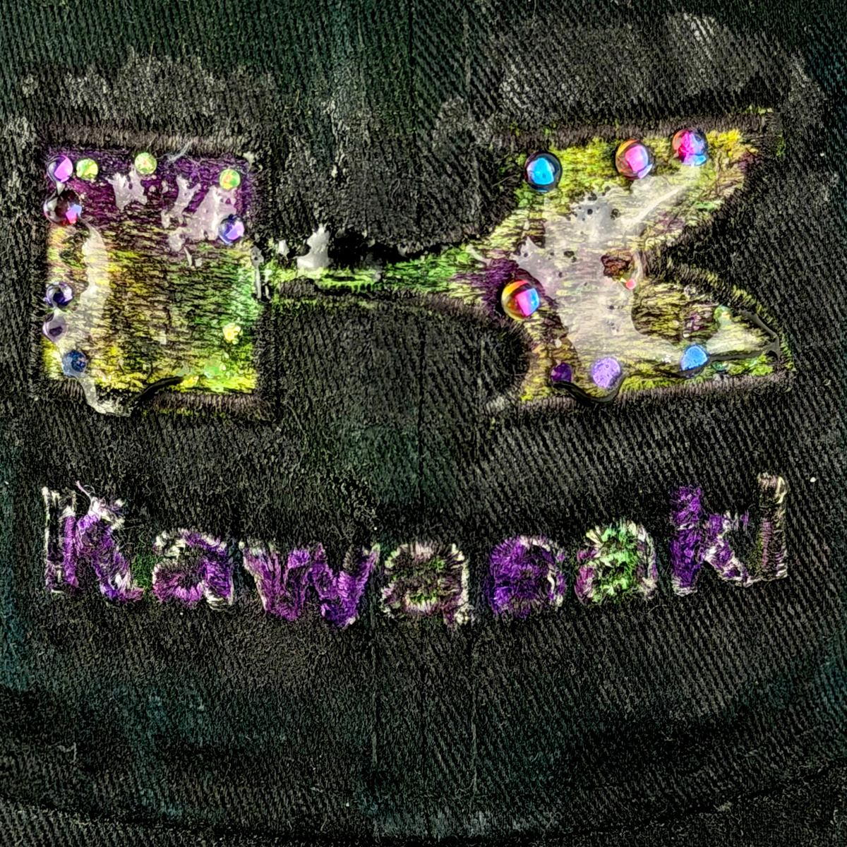 Close-up of a dark green and black logo featuring a distressed patch with a stylized 'K' and the word 'Kawasaki' in purple embroidered letters.