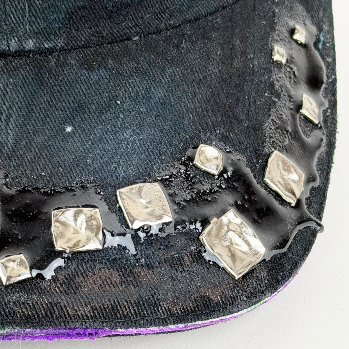 Close-up of a black fabric cap with metallic silver foil square accents arranged along a curved seam, with glossy wet highlights reflecting off the surface.