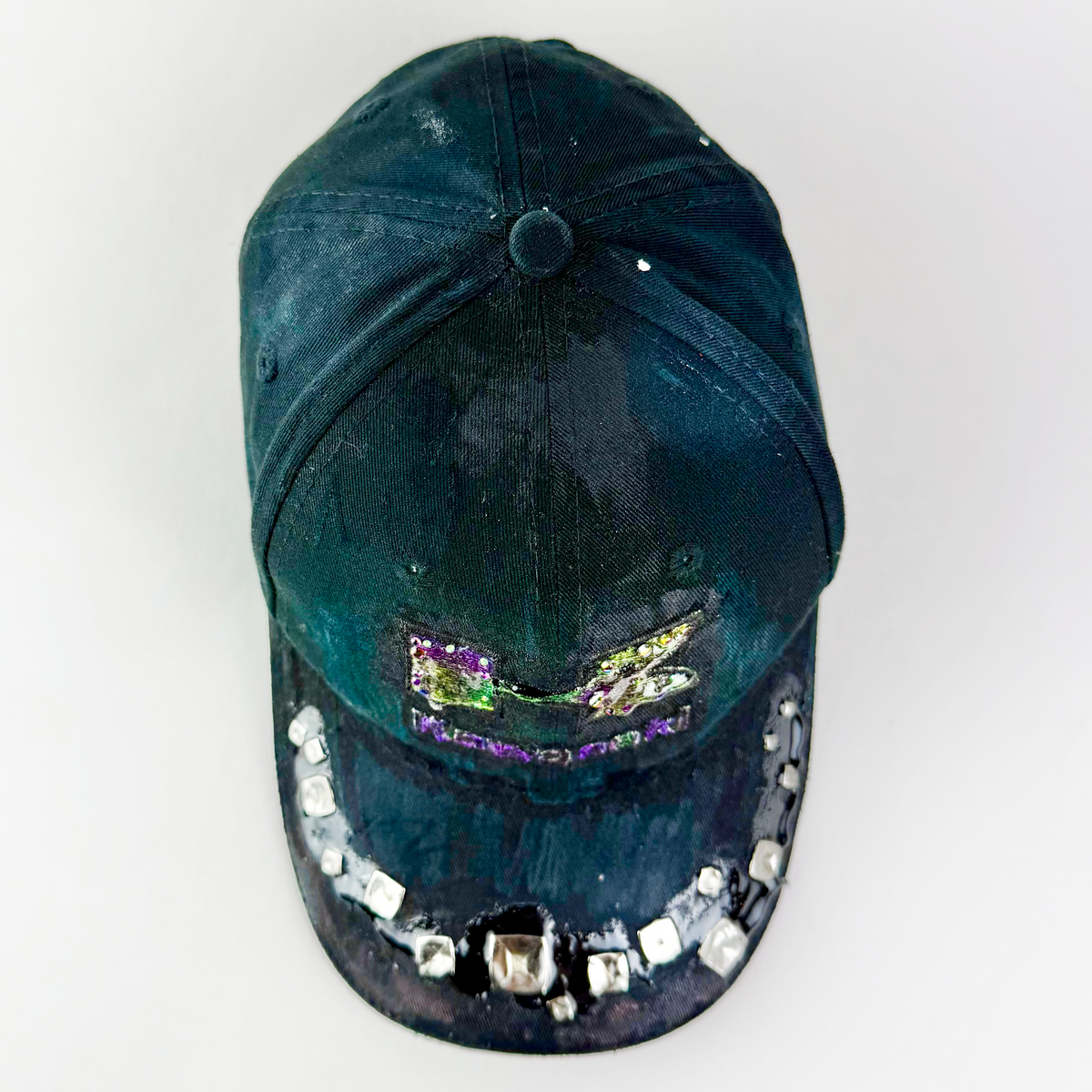 A dark teal-green distressed baseball cap with a colorful embroidered patch on the front and a row of silver rhinestones along the brim, shown against a plain white background.