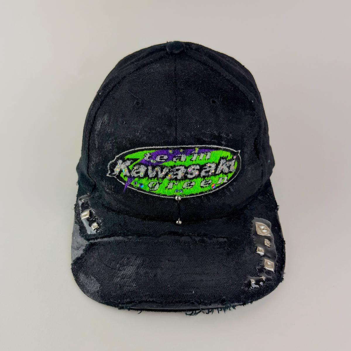 ASTROBOI* x Kawasaki 'Faust' black green and purple rhinestone studded cap with rhinestone-embellished Kawasaki logo, stainless steel piercing, and deconstructed visor, front view.