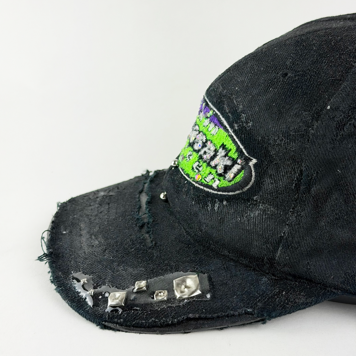 A close-up of a distressed black baseball cap with a green and purple embroidered patch on the side and silver studs on the brim, set against a plain white background.