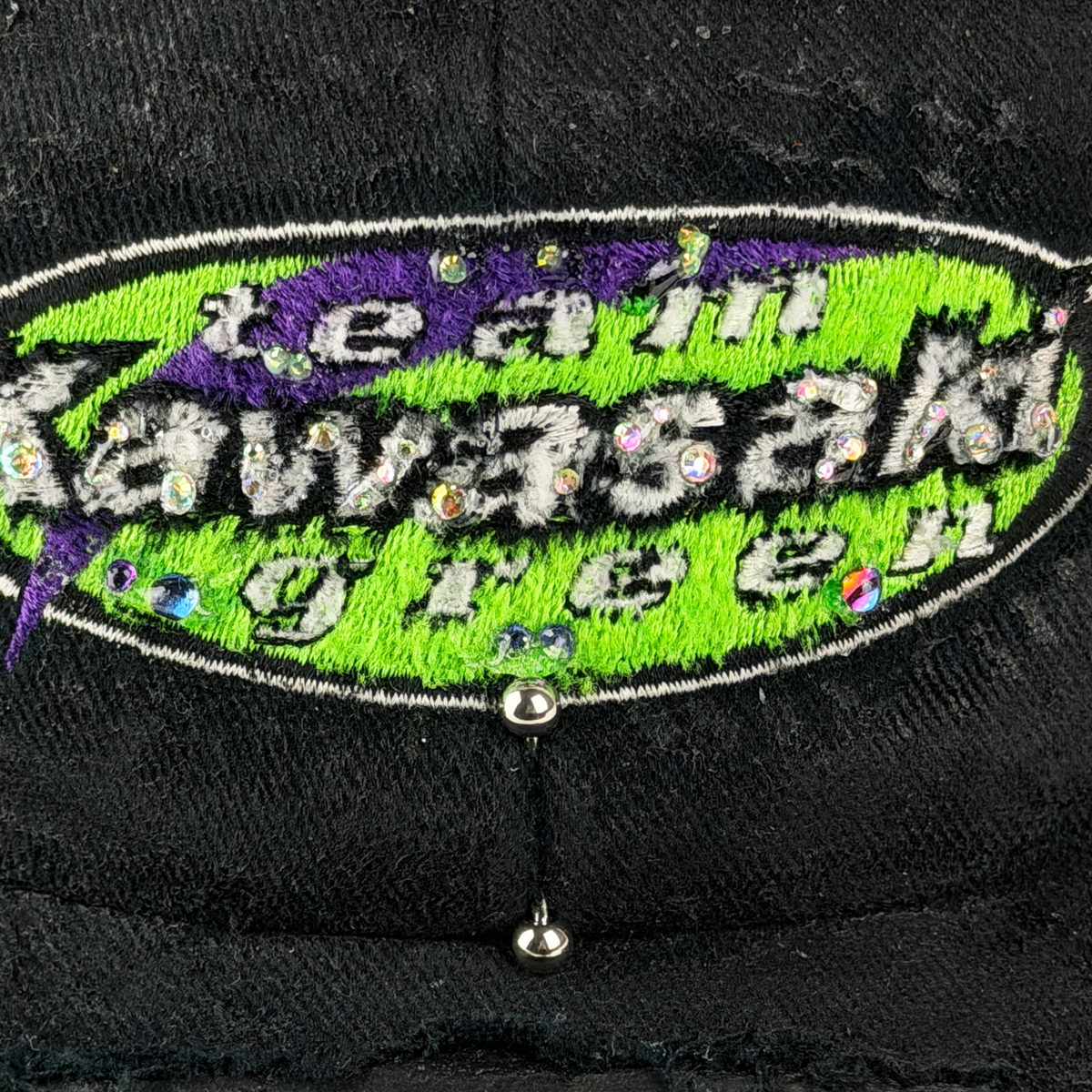 Close-up of a black embroidered patch reading 'Team Kawasaki Green' with purple and neon green accents, rhinestone details, and a silver metal stud hanging below.