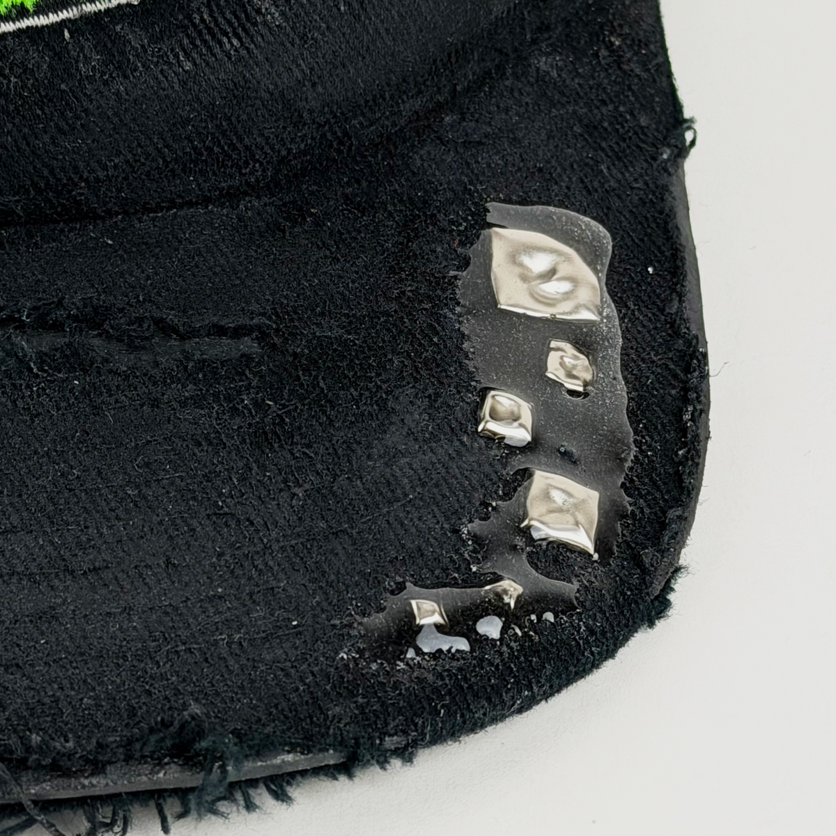Close-up of a distressed black baseball cap with frayed edges, a small green embroidered patch, and several silver rhinestone studs on the brim, photographed on a white background.