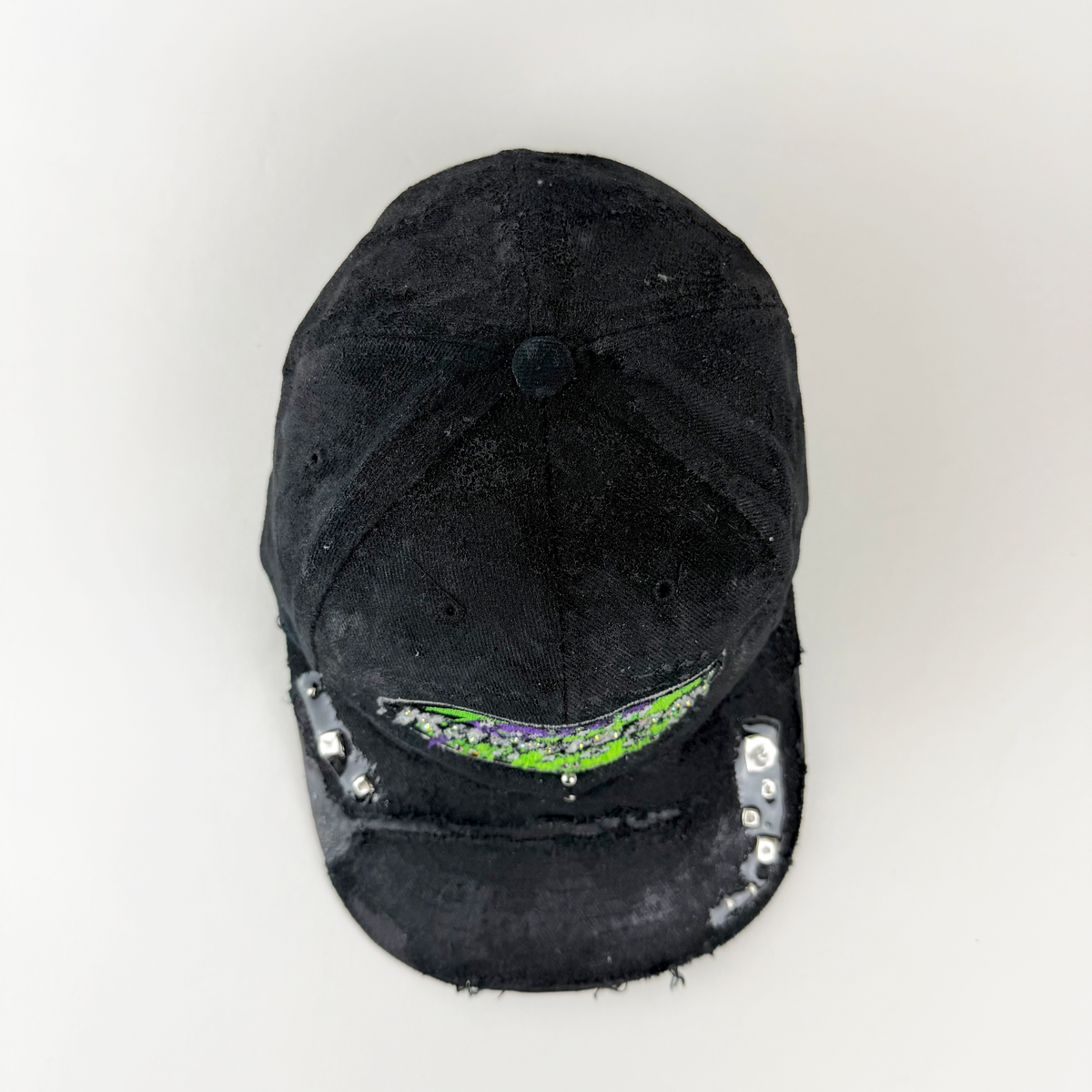 A distressed black baseball cap with a green and black graphic on the front and silver sequins along the brim, photographed against a plain white background.
