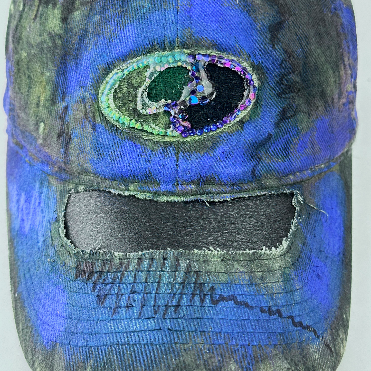 Close-up of ASTROBOI* x Mossy Oak 'Ultraviolet' showing rhinestone-embellished Mossy Oak logo and ultraviolet flowing paint treatment on Real Tree camouflage fabric.