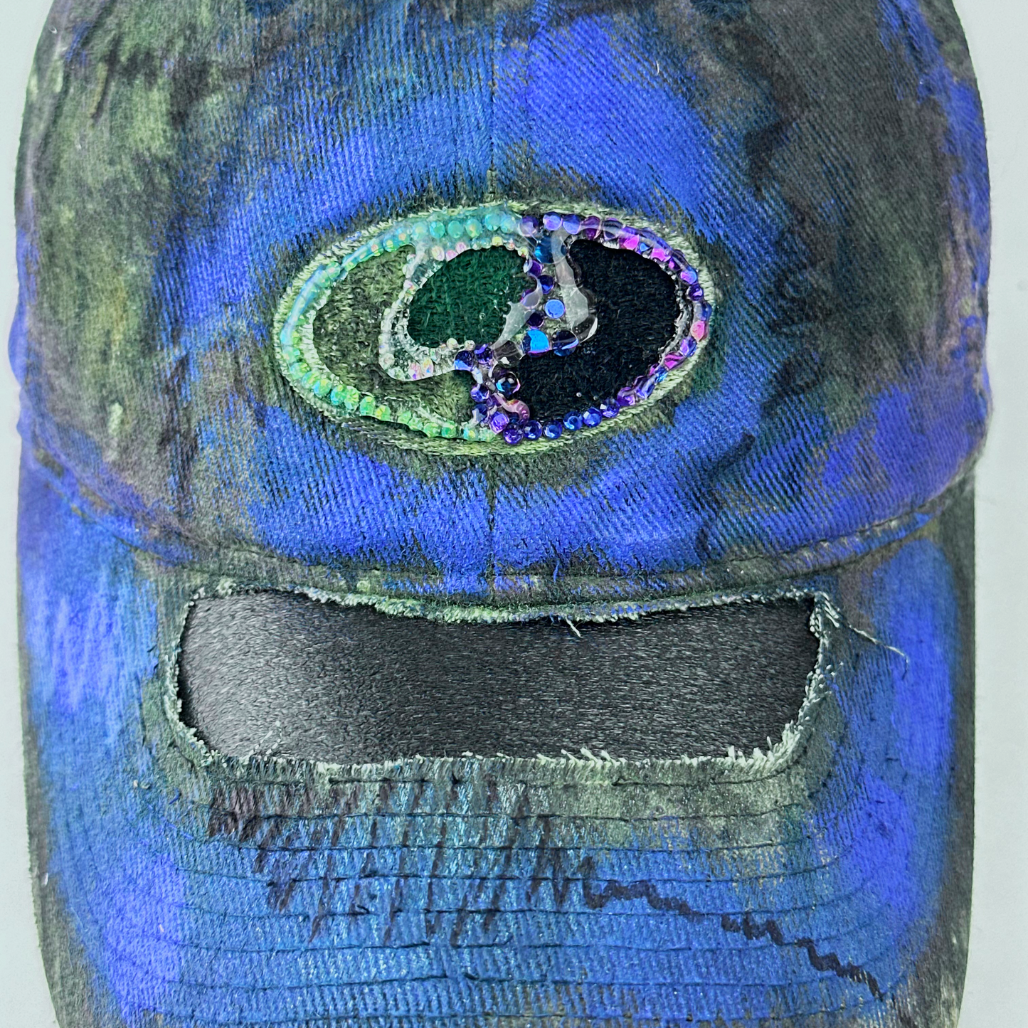 ASTROBOI* x Mossy Oak 'Ultraviolet' brown and purple Real Tree camouflage hat with rhinestone-embellished Mossy Oak logo at front, ultraviolet flowing paint treatment, and deconstructed visor, logo close up.