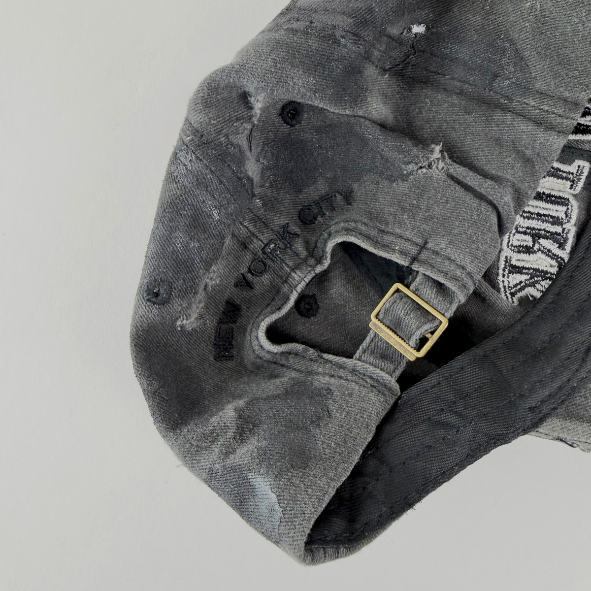 Close-up of a distressed gray washed baseball cap with black embroidered text reading 'NEW YORK CITY' and a gold-tone buckle on the strap, shown against a light gray background.