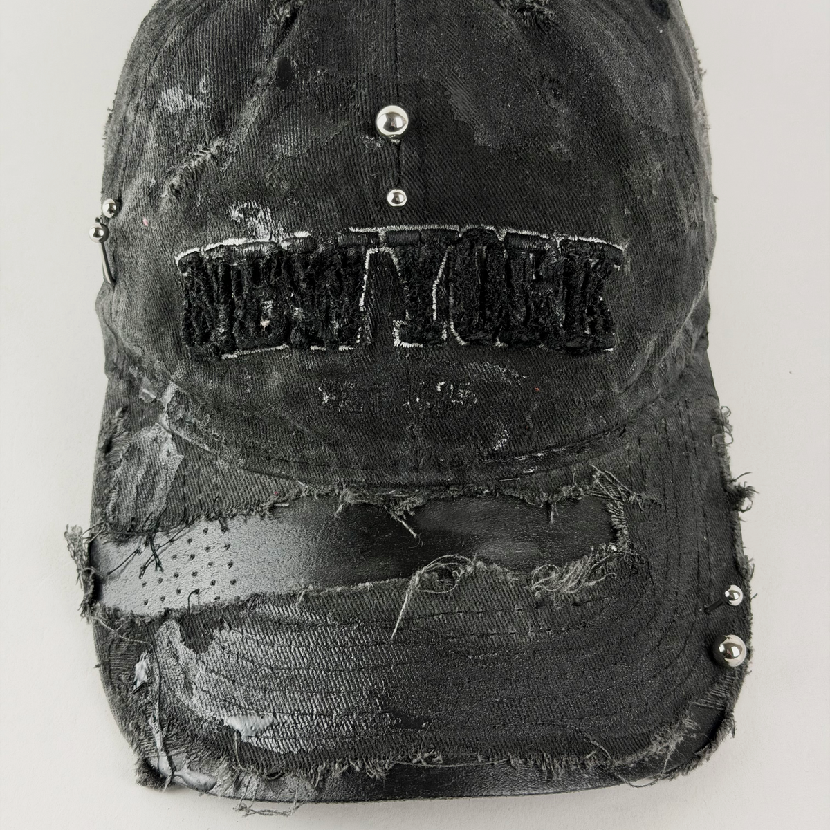 A distressed black baseball cap with a heavily ripped and frayed texture, featuring a sewn-on 'NEW YORK' patch on the front and silver metal studs.