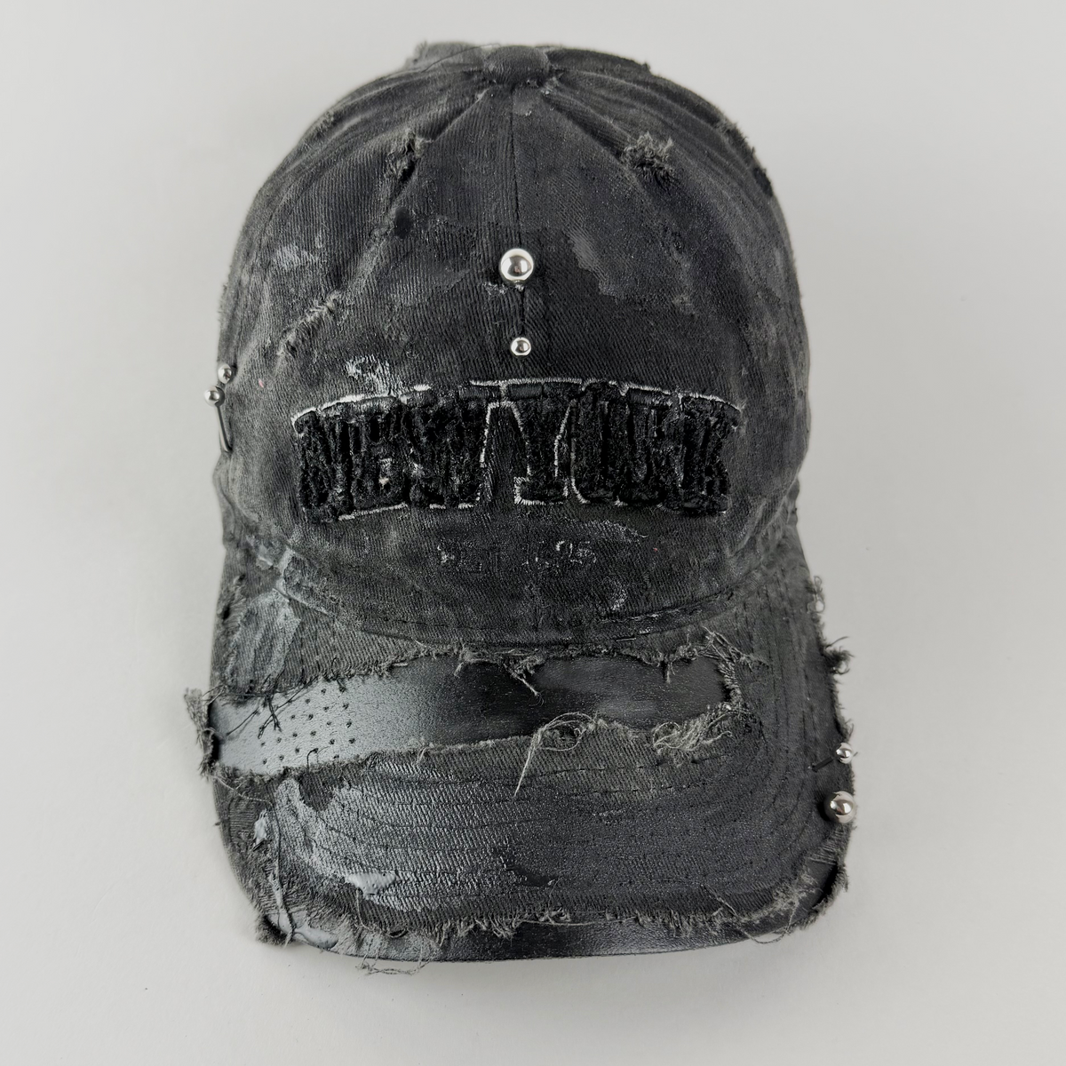 Close-up of ASTROBOI* 'New York Grunge' showing 'New York' embroidered logo, black paint detail, and stainless steel piercing on distressed black fabric.
