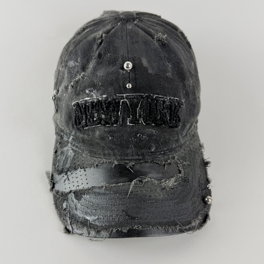 Close-up of ASTROBOI* 'New York Grunge' showing 'New York' embroidered logo, black paint detail, and stainless steel piercing on distressed black fabric.