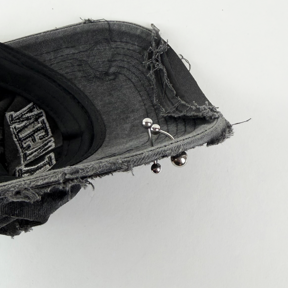 A close-up of a distressed black denim cap held by a hand, showing a silver double-barbell nose ring worn on the brim.