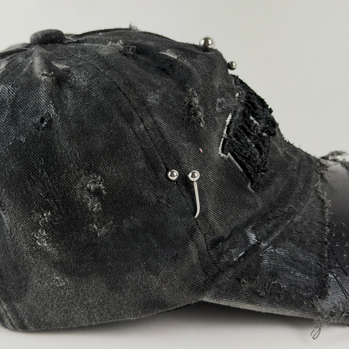 A close-up of a distressed black denim baseball cap with ripped details, frayed edges, and silver metal studs and barbell embellishments on the front.